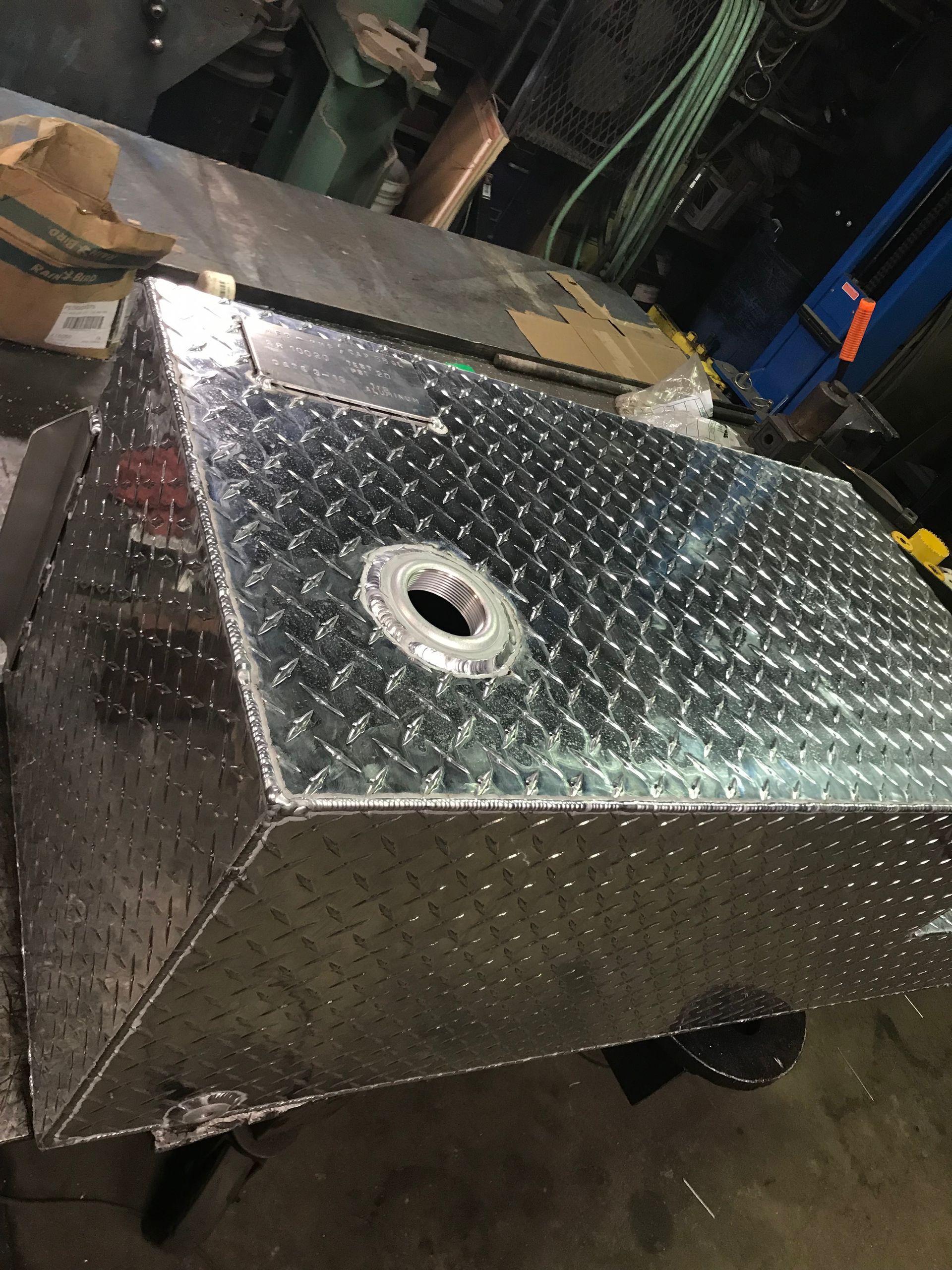 A rectangular diamond plate aluminum storage tank with a central circular opening, shown in a workshop setting.