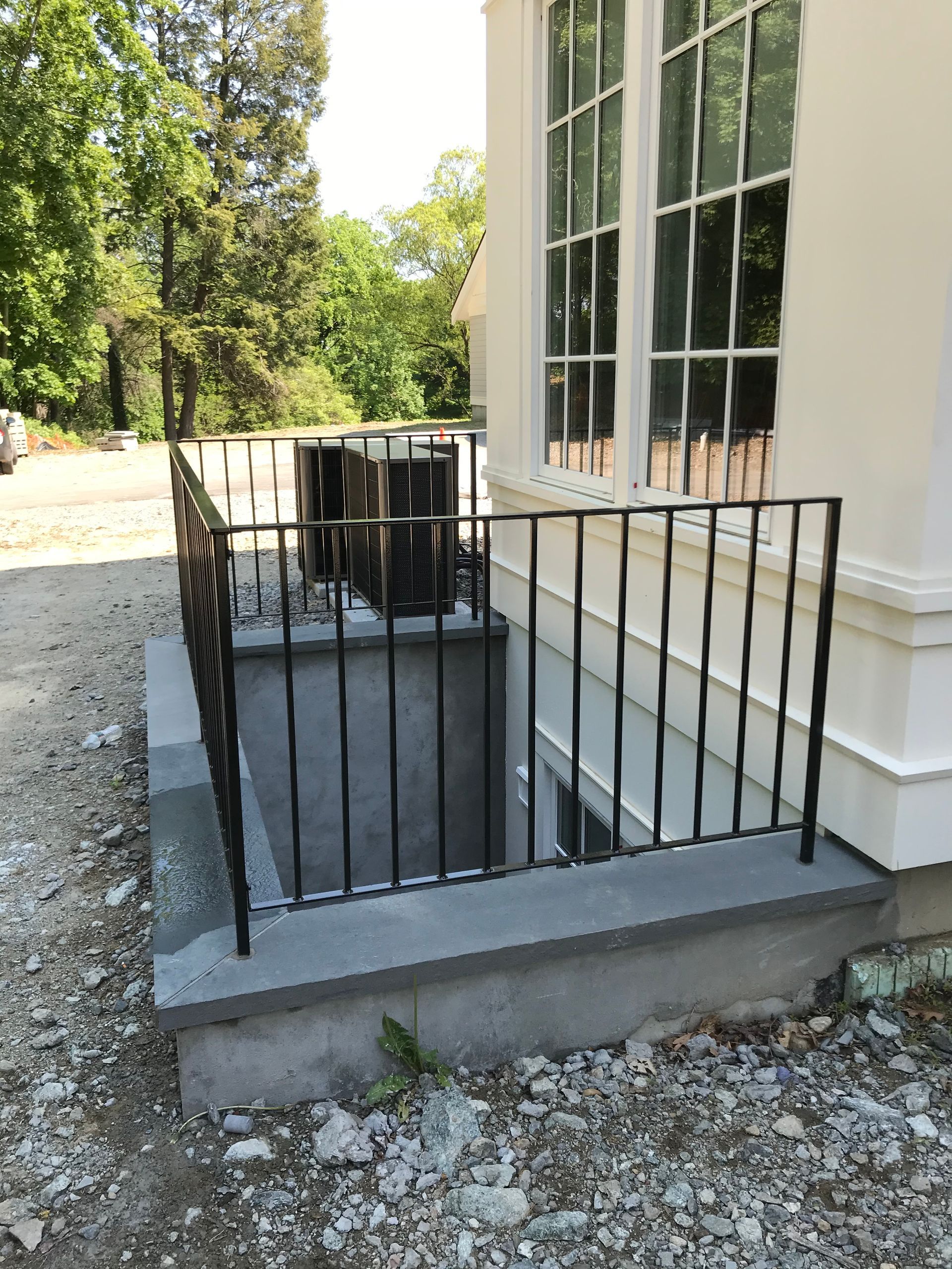 A metal railing encloses a concrete basement stairwell next to a house with white siding and a large window.
