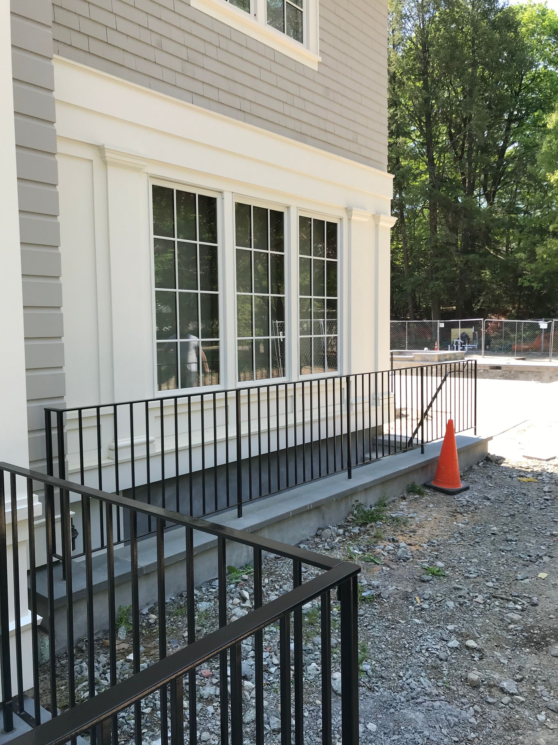 A grey and white house exterior with a black metal railing protecting a raised foundation near a gravel path and orange cone.