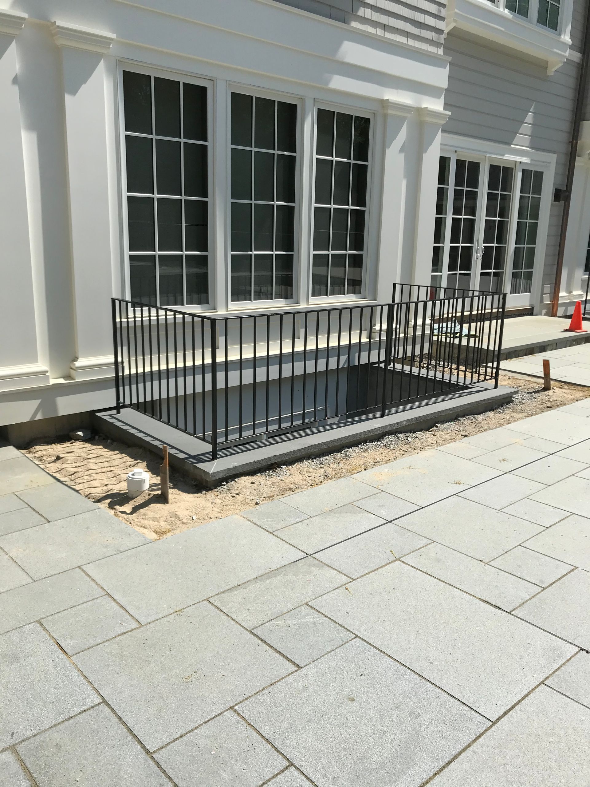 A basement window well with a black metal railing set in a stone patio area next to a building.