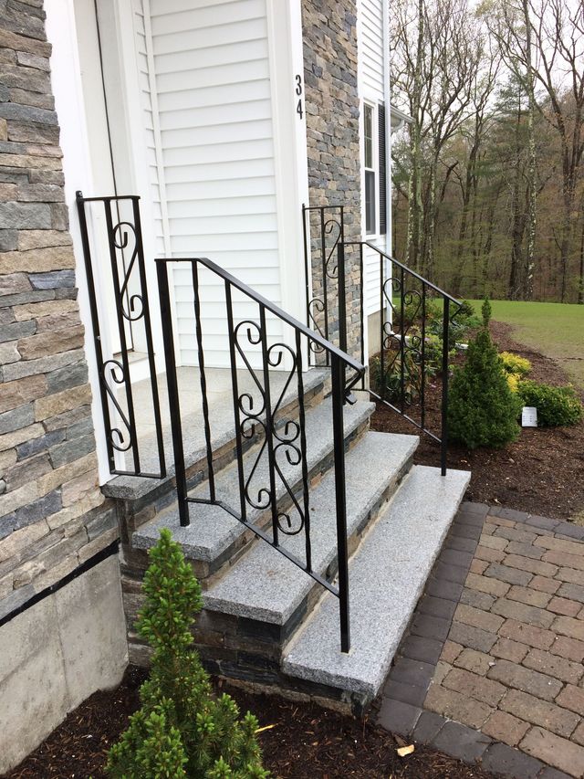 Black wrought iron handrails on light gray stone steps leading to a house entrance with stone siding.