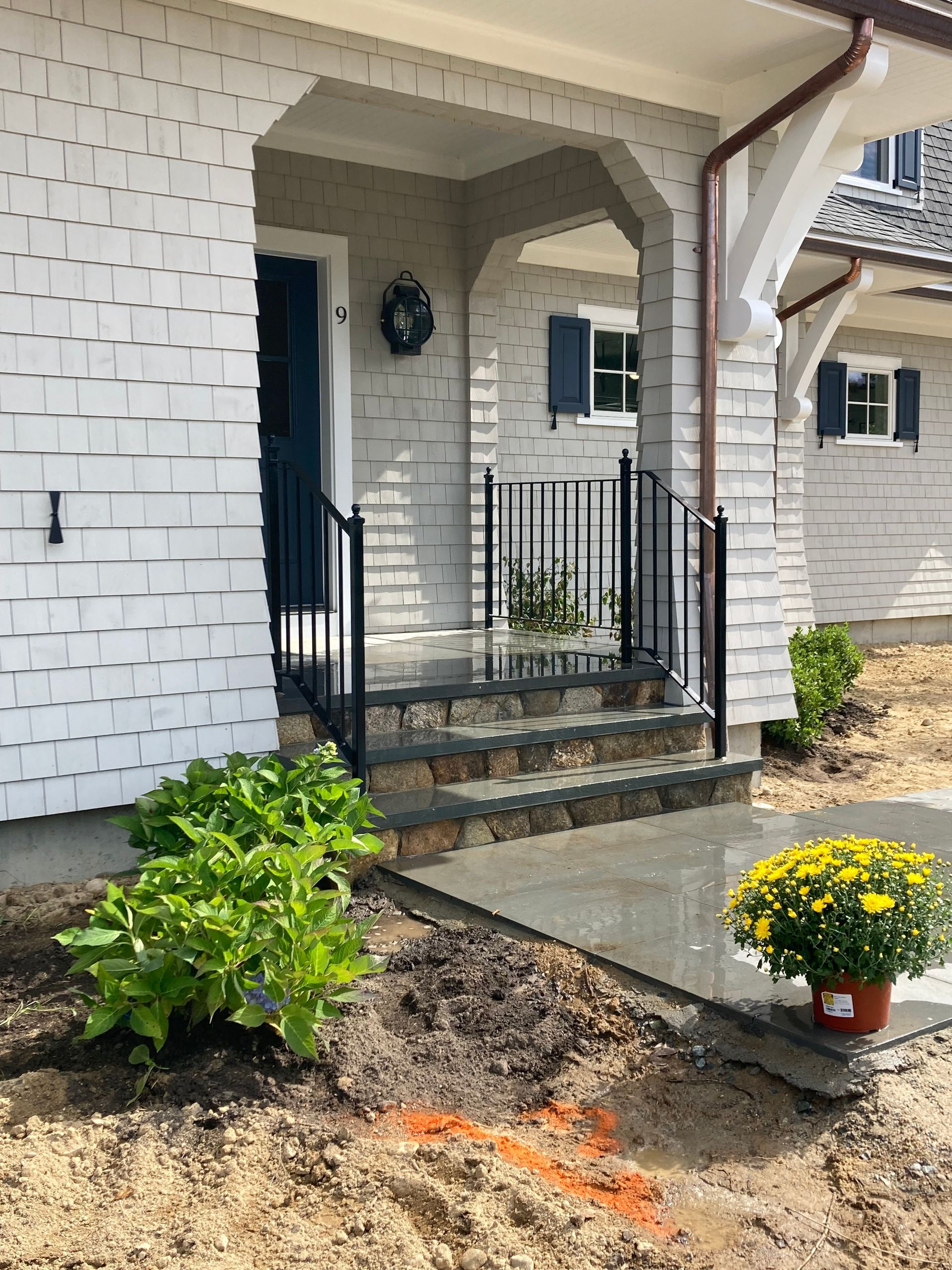 A white shingled house entrance with a stone-stepped porch, black metal railings, and a potted yellow mum in front.