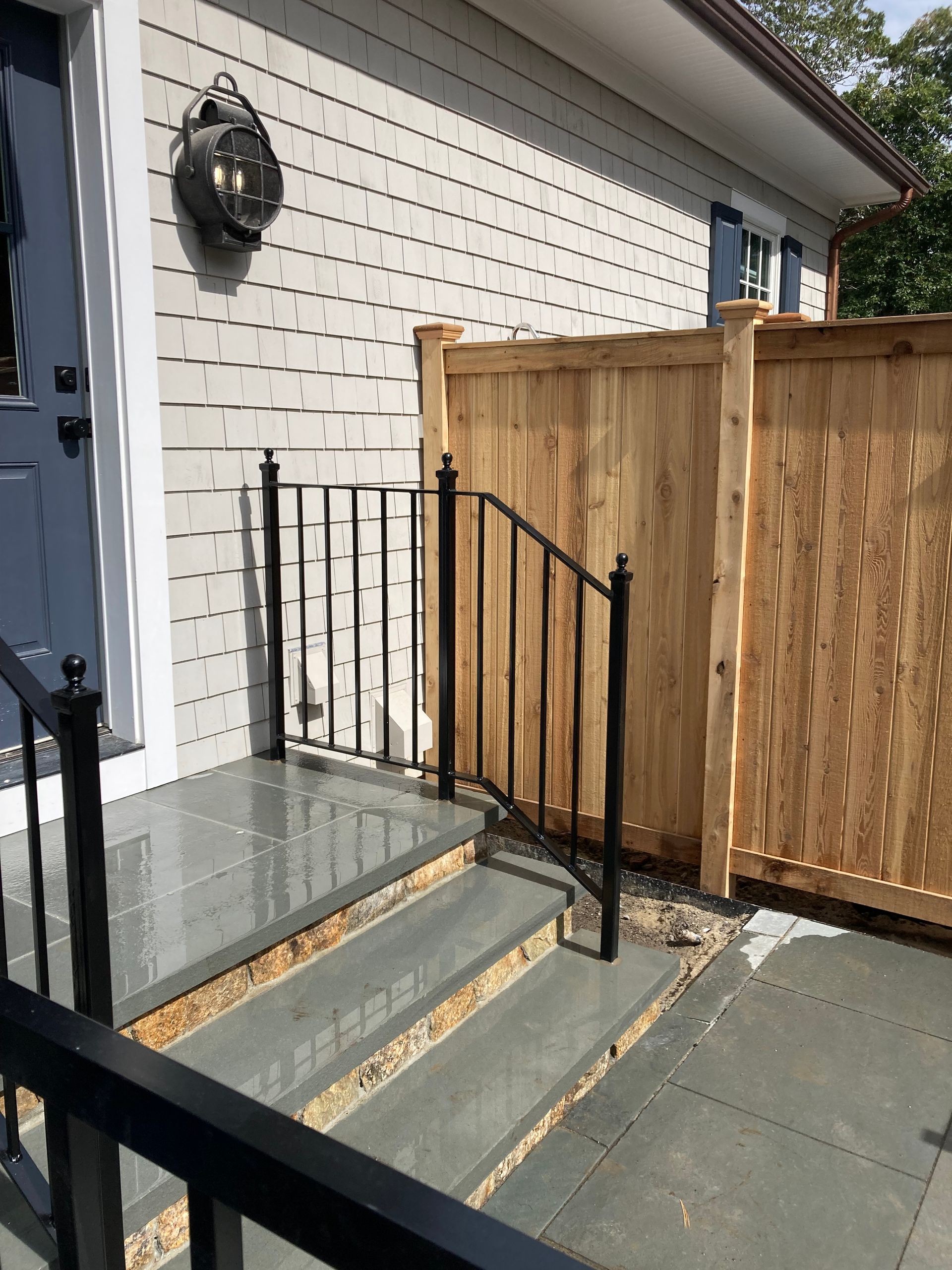 A blue door and grey stone steps with black metal railings next to a wooden fence on the side of a light-colored building.