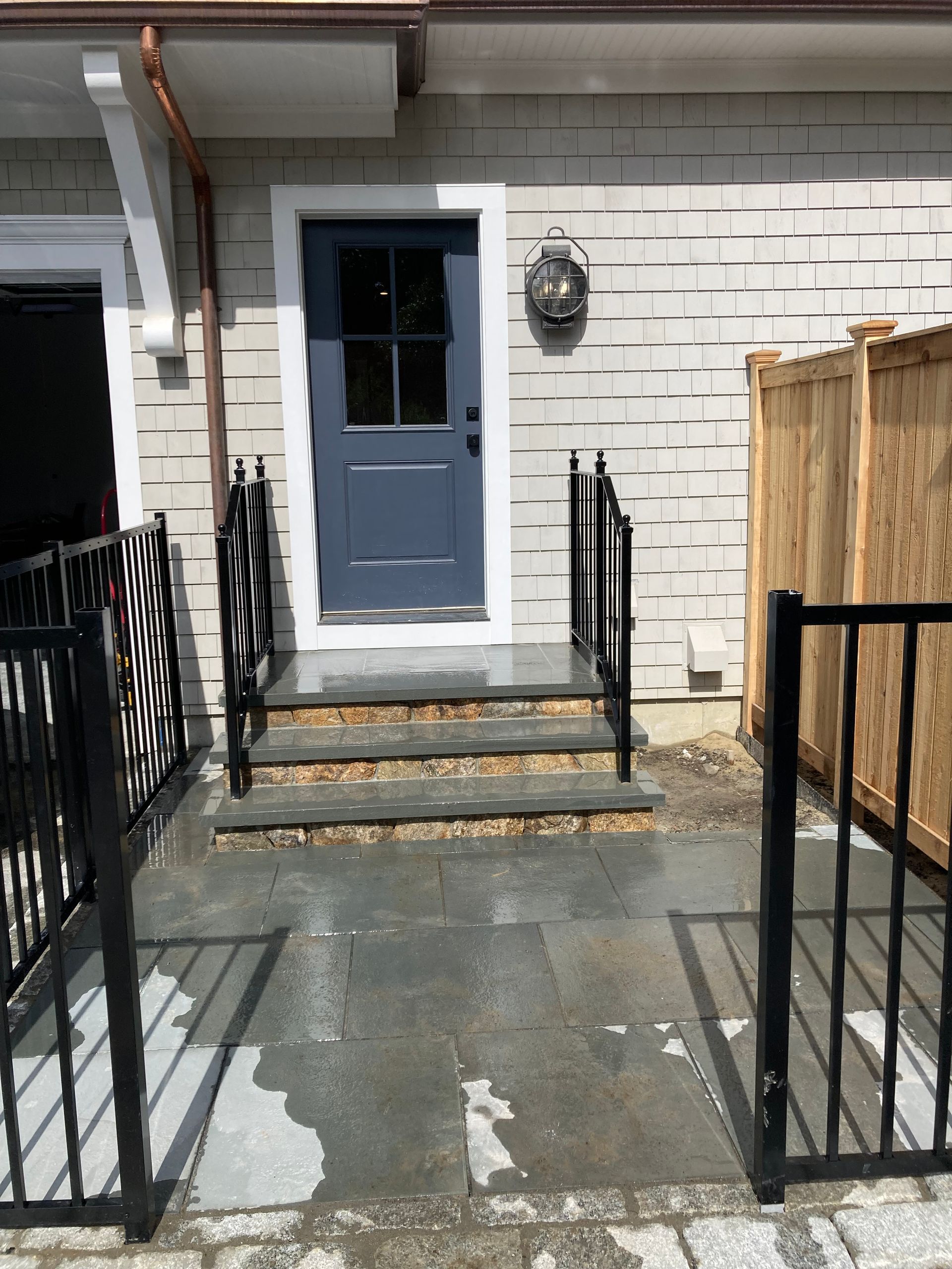 A blue front door with a window leads to three grey stone steps, framed by black railings and a light-colored wood fence.