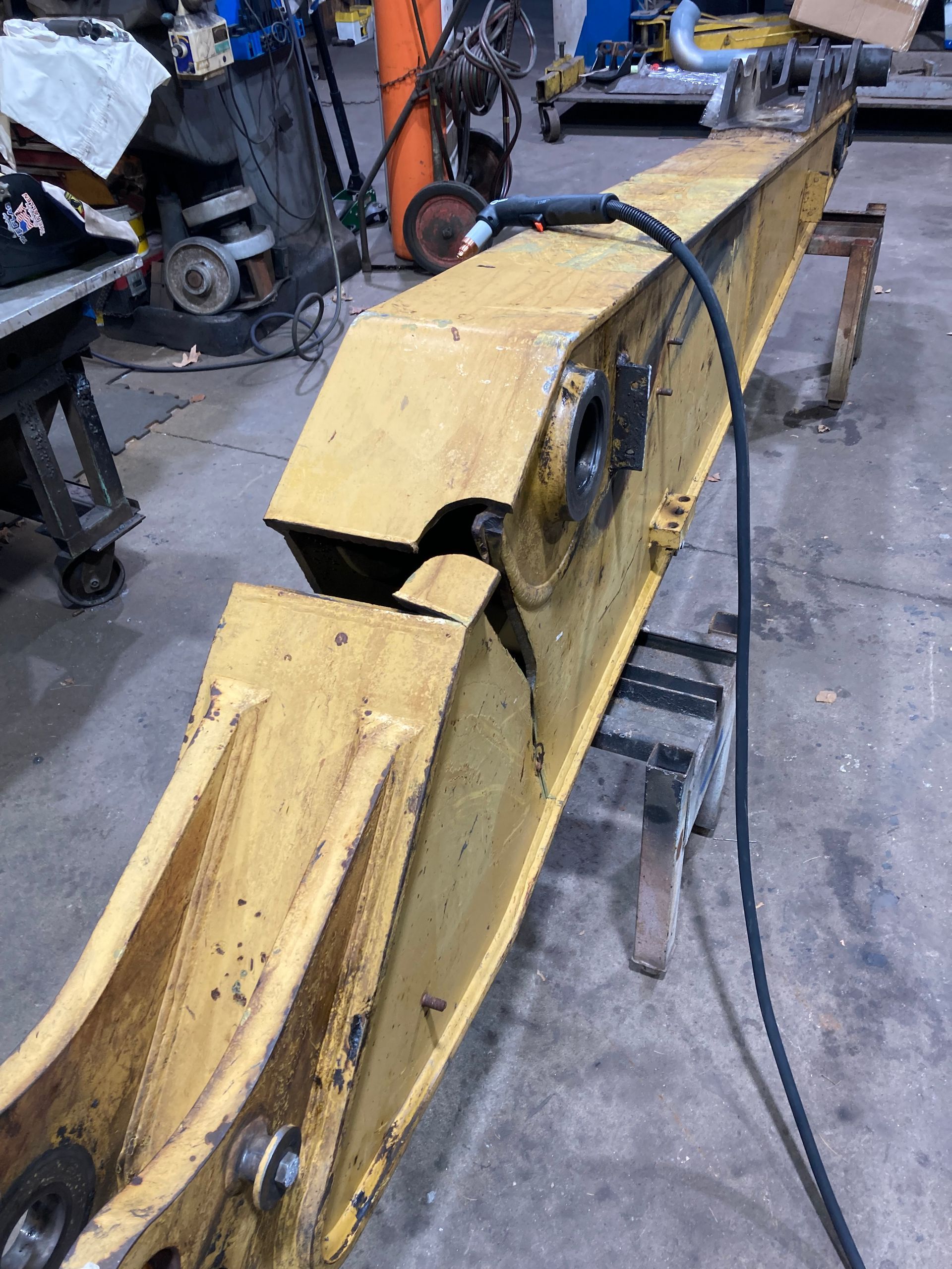 A large, yellow, damaged excavator boom rests on metal supports in a workshop, showing a clear, jagged structural crack.
