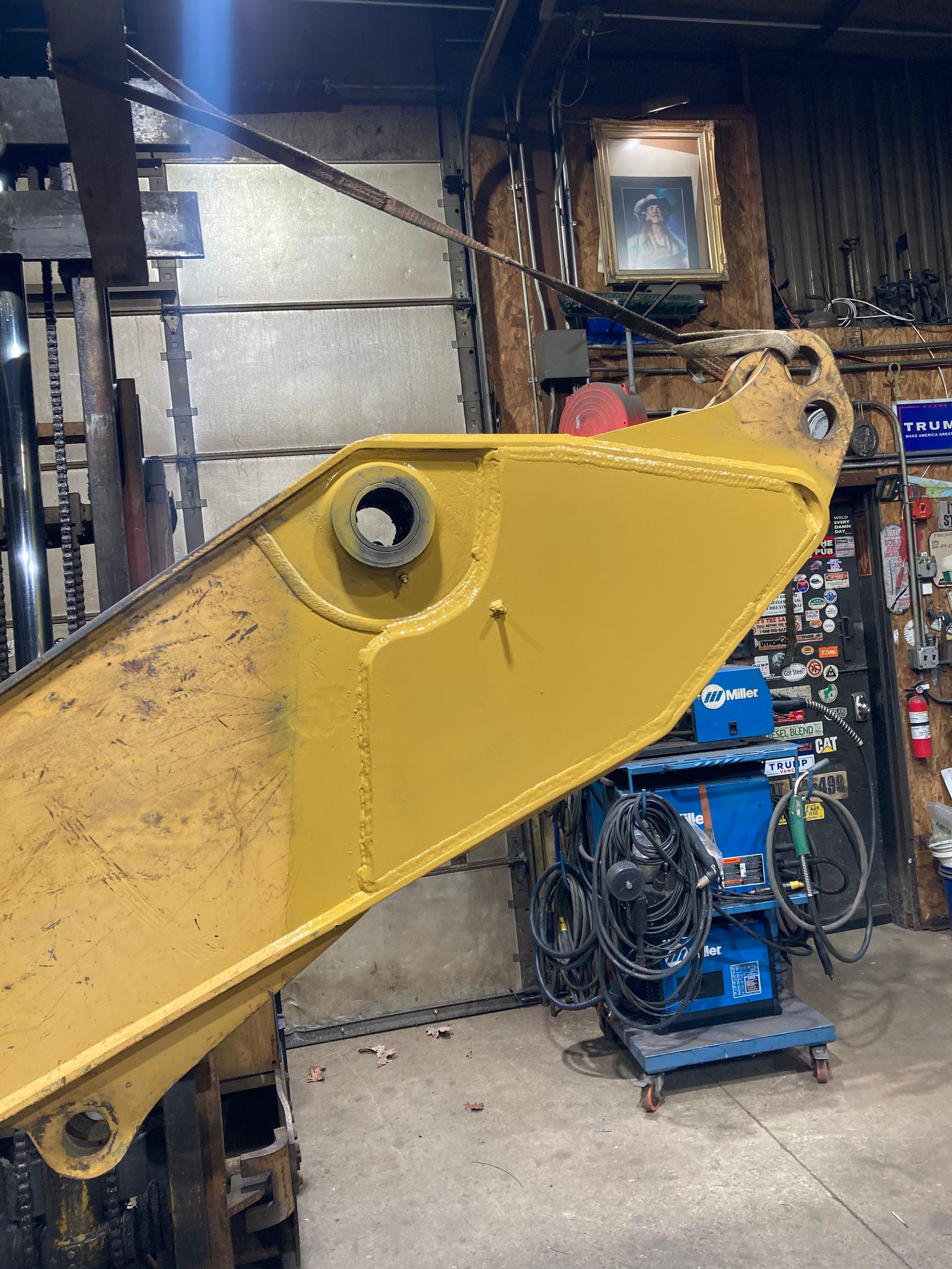 A yellow industrial excavator boom arm is suspended in a workshop, showcasing a large metal pivot hole and welded frame.