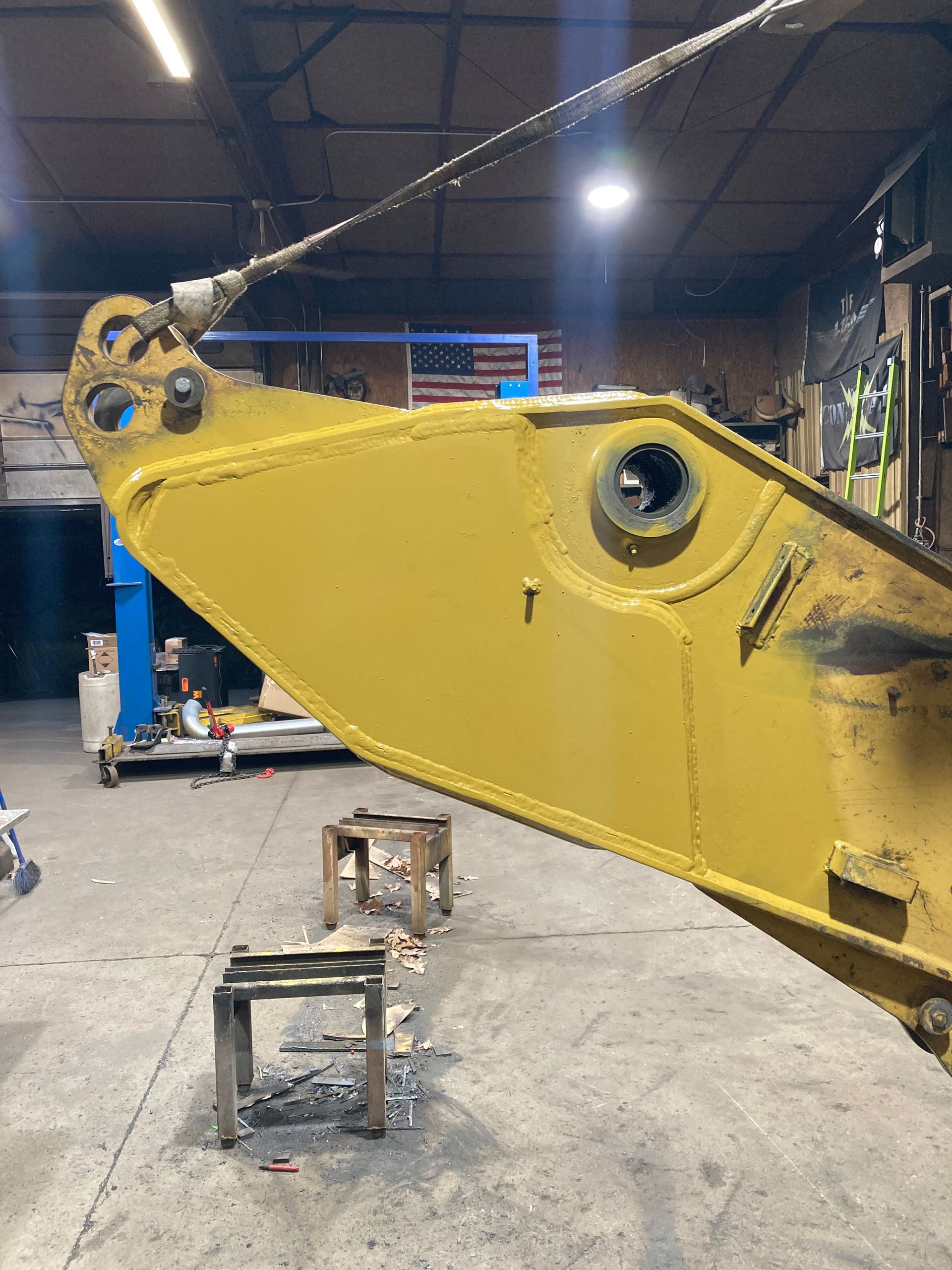 A yellow excavator boom component hangs in a workshop, suspended by a cable near an open bearing housing.