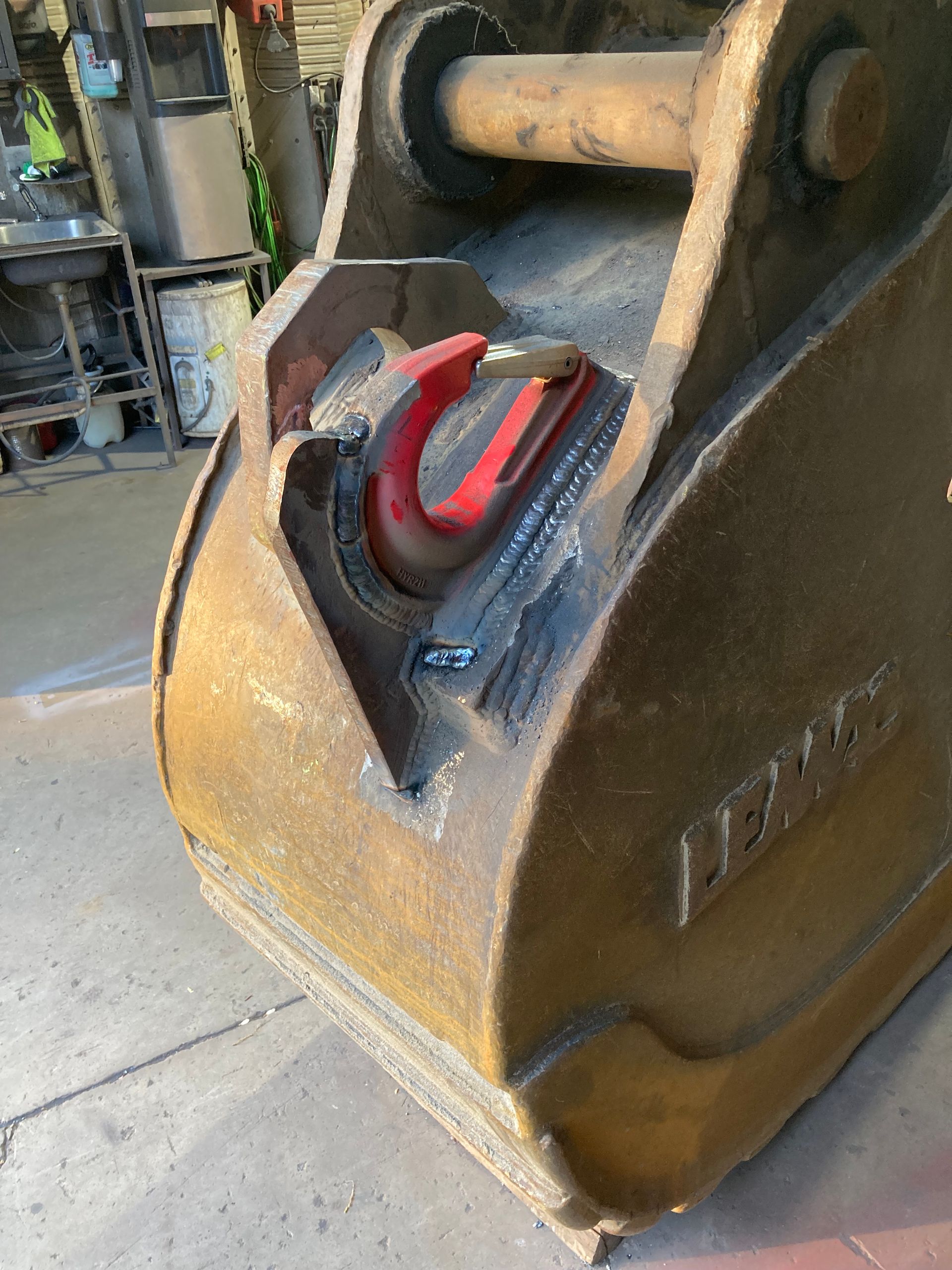 A close-up view of a metal excavator bucket featuring a welded red lifting hook, set in an industrial workshop.