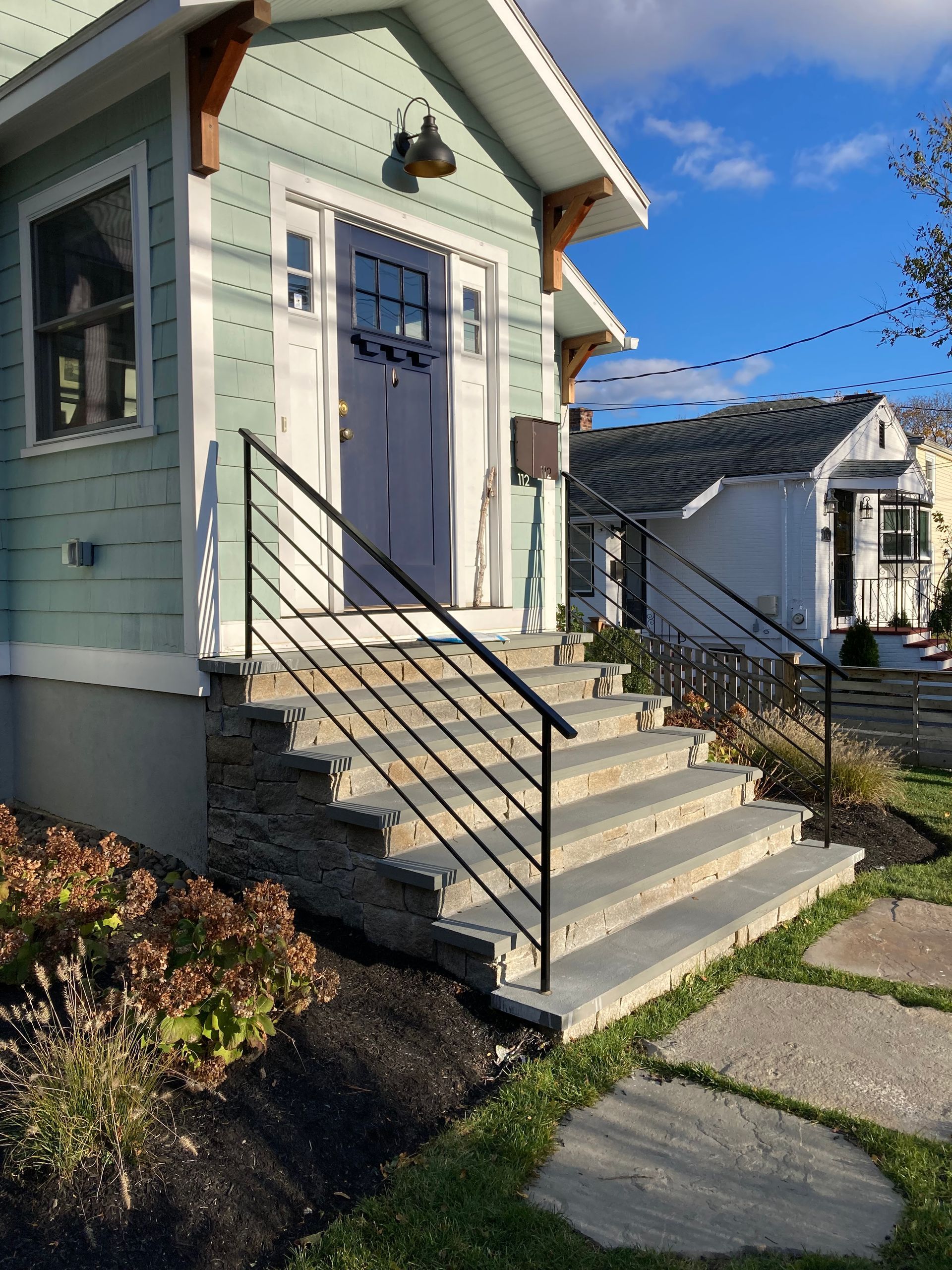 A light green house with a purple front door, black railings, and stone steps, viewed on a sunny day.