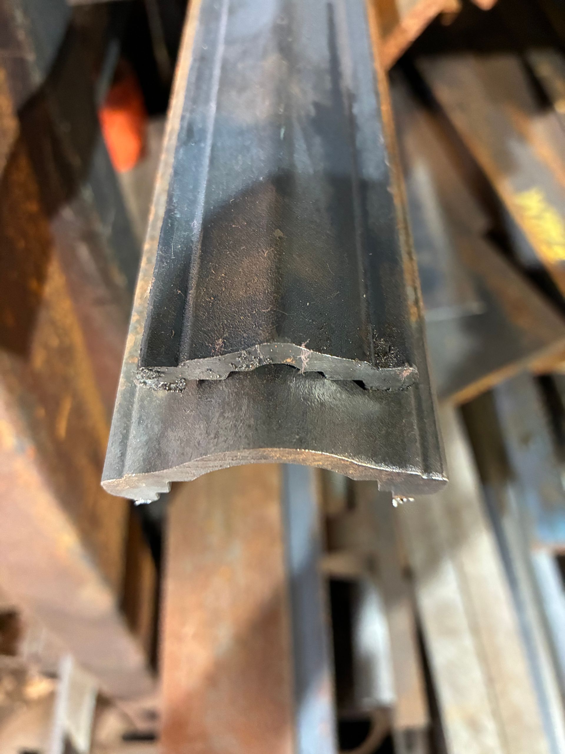 A close-up of a dark metal T-shaped trim or molding piece with a curved underside, resting in a cluttered workshop.