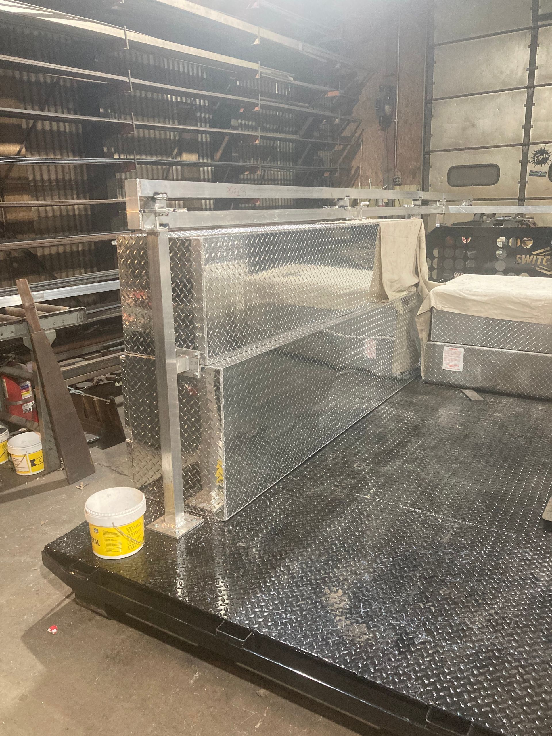 Metal workbench with a diamond-plate surface and raised shelving in a workshop with shelves of supplies in the background.