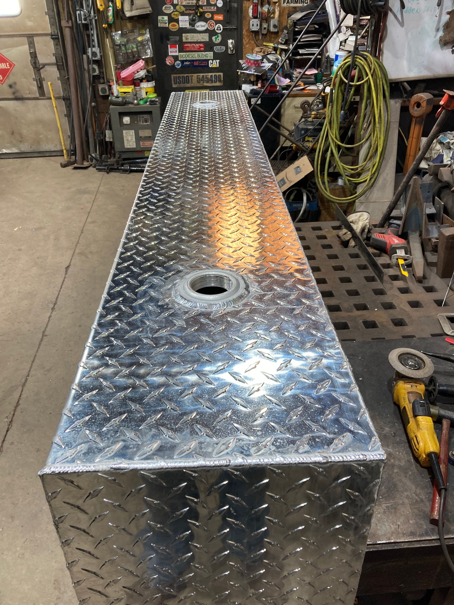 A long, rectangular diamond-plate aluminum fuel tank with a circular fill neck sitting on a metal workbench in a workshop.