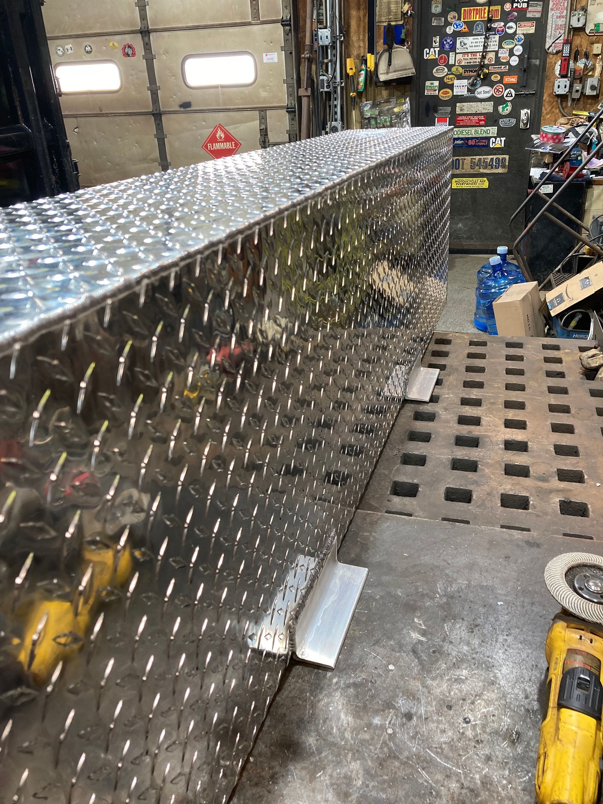 A long, rectangular diamond-plate aluminum storage box sits on a metal workbench in a cluttered workshop.