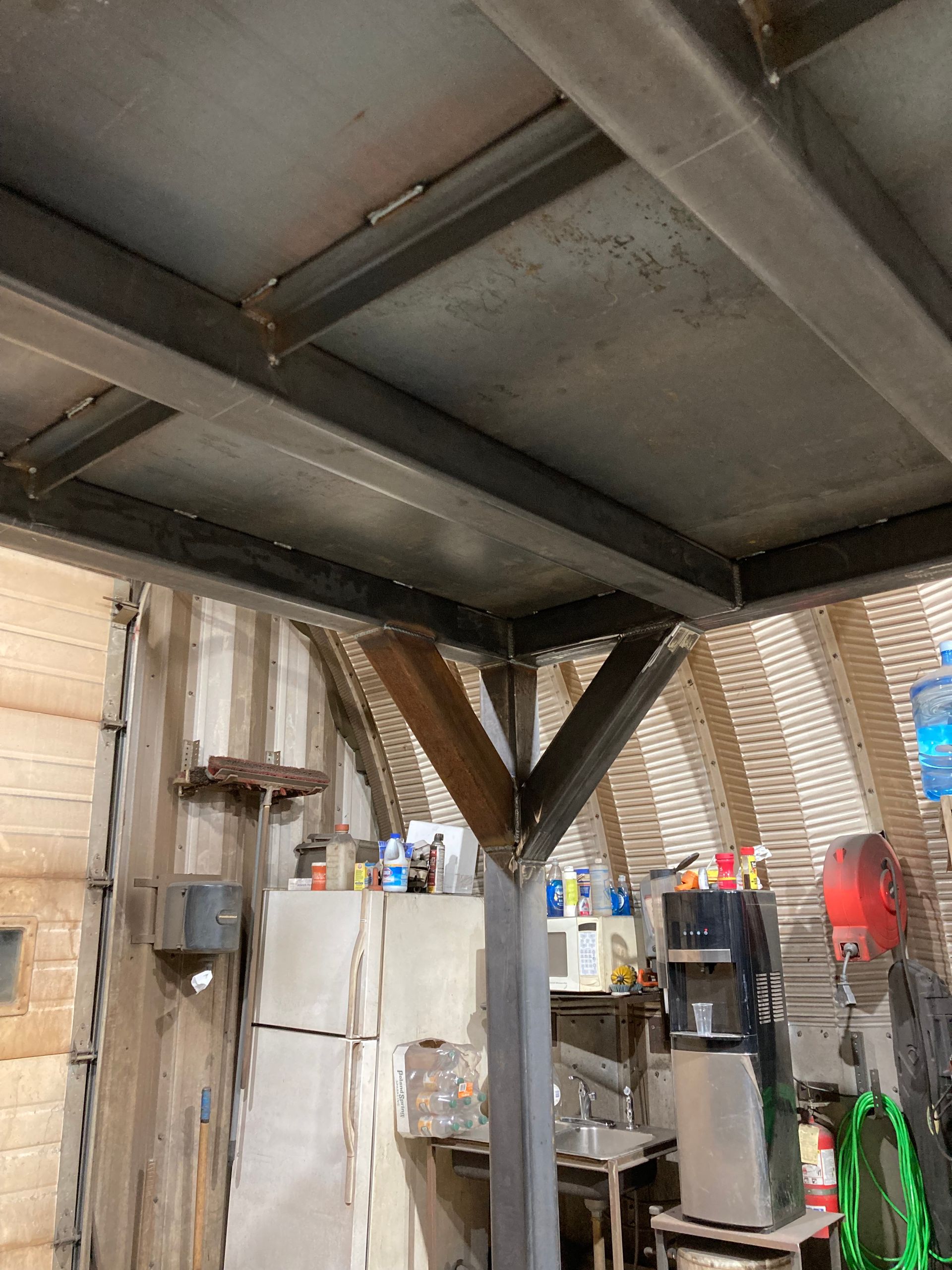 Steel support column with angled braces under a metal framed loft inside a corrugated metal workshop.