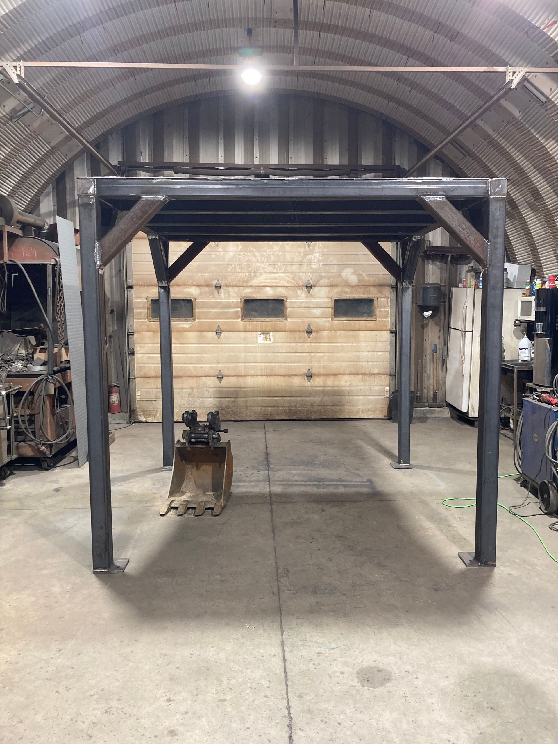 A steel rectangular frame structure standing in a workshop, featuring four vertical columns and reinforced corner braces.