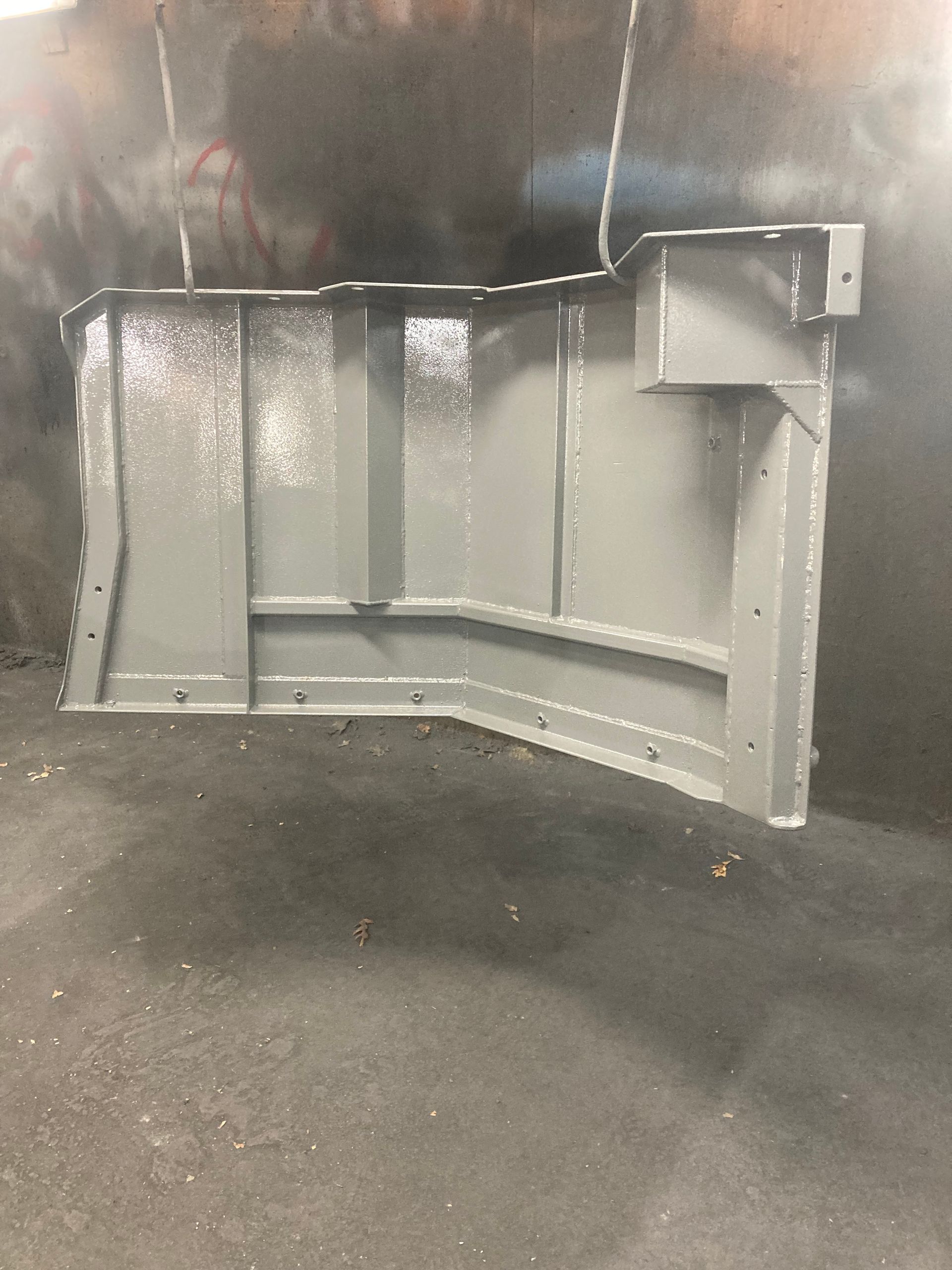 A metal component with structural ribs, painted in light grey, hanging in an industrial workshop setting.