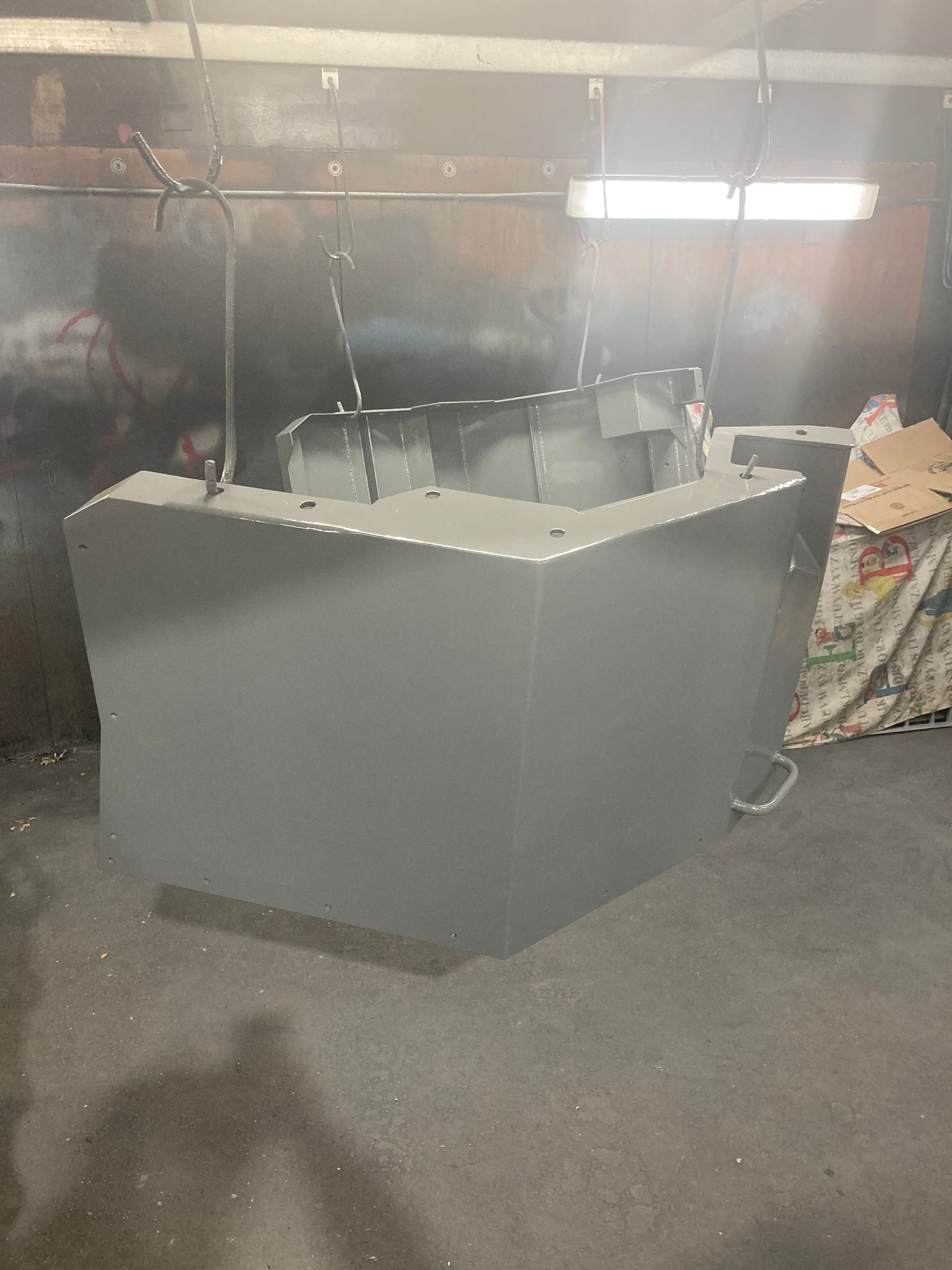 A gray, L-shaped metal structure hangs from chains inside a workshop or industrial spray booth.