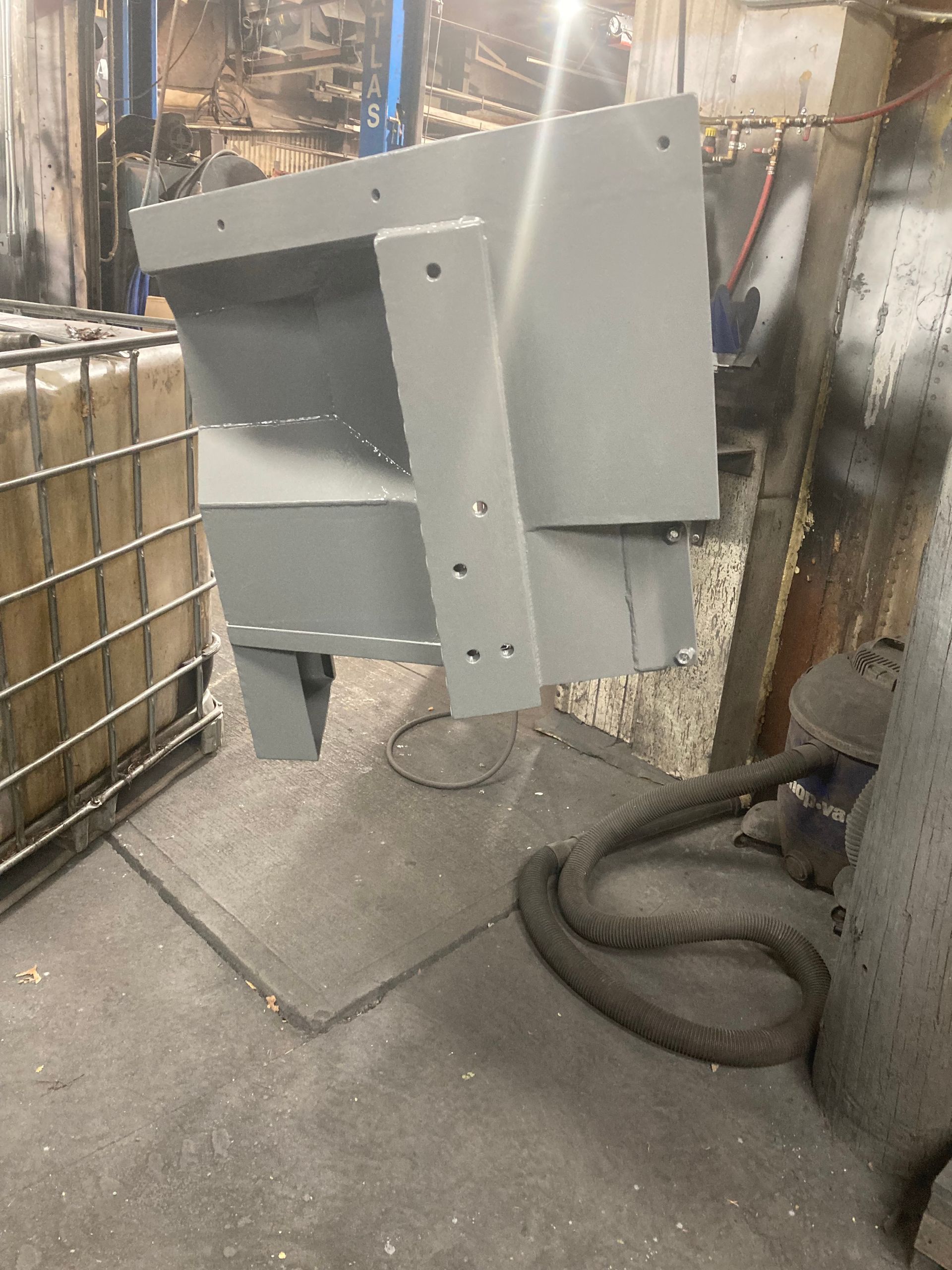 A light gray industrial metal part hangs in a workshop, featuring a flat mounting bracket with several pre-drilled holes.