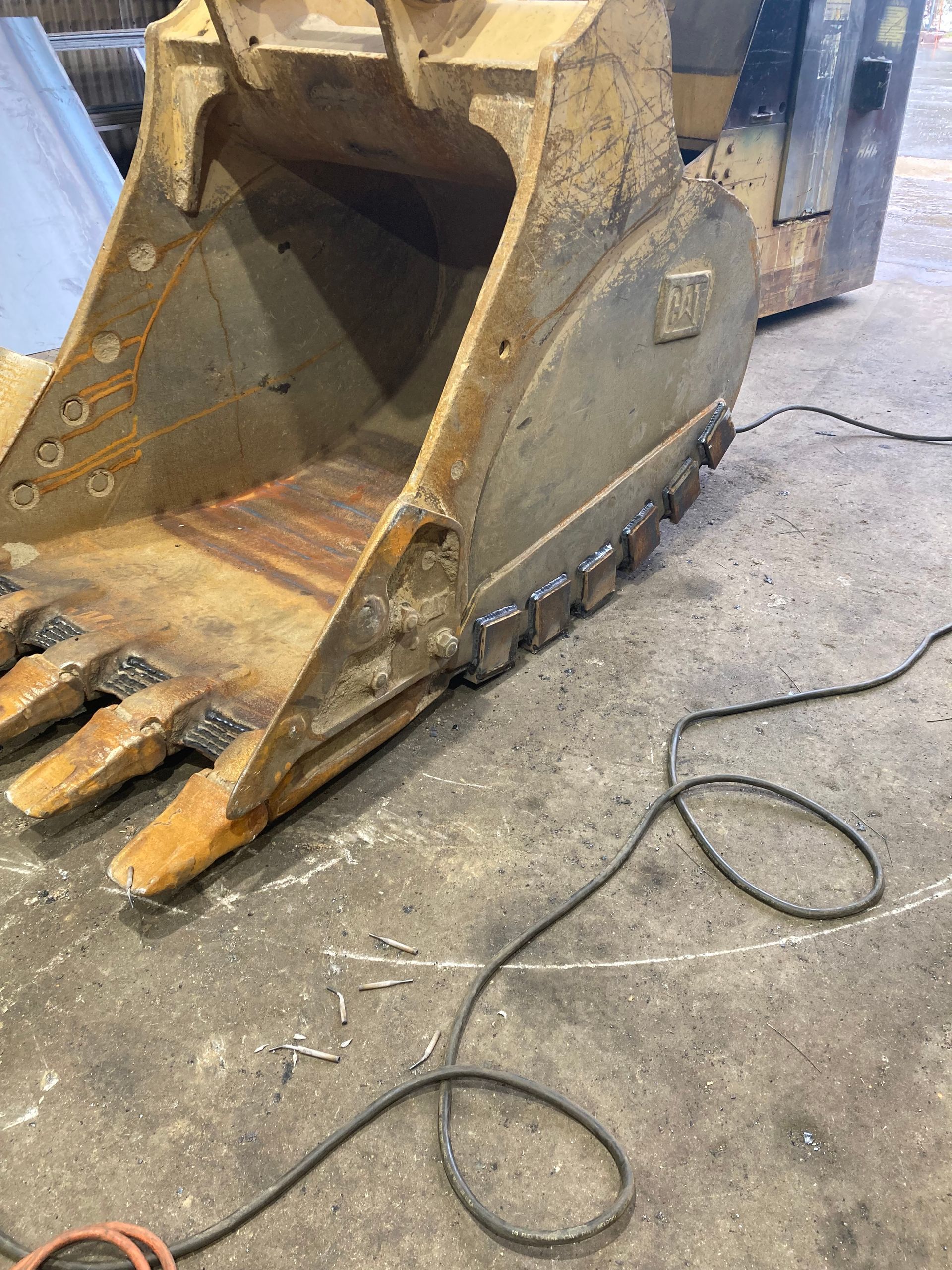 A yellow Caterpillar excavator bucket sits on a concrete shop floor, showing signs of heavy use and welding repairs.
