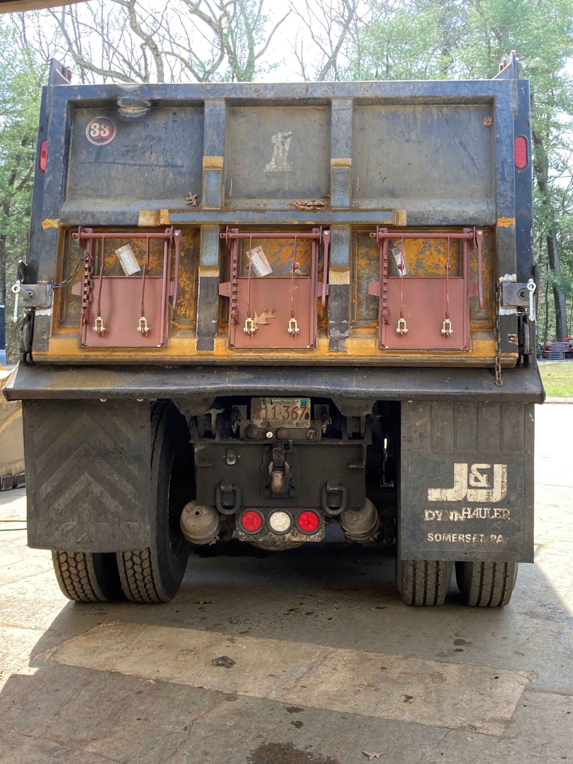 The rear view of a dark metal dump truck with three rusted access panels on the tailgate, parked on a paved surface.