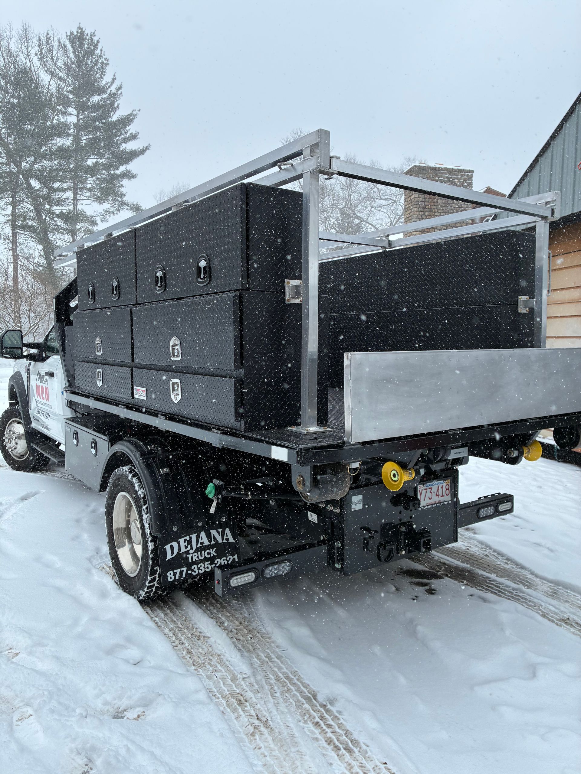 A flatbed truck with black storage boxes and a metal rack, parked in a snowy setting.