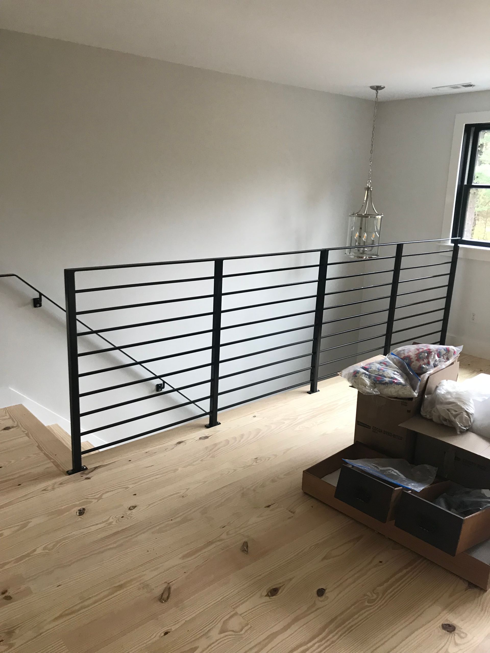 A modern black metal horizontal railing lines an indoor staircase and landing with light-toned wooden floorboards.