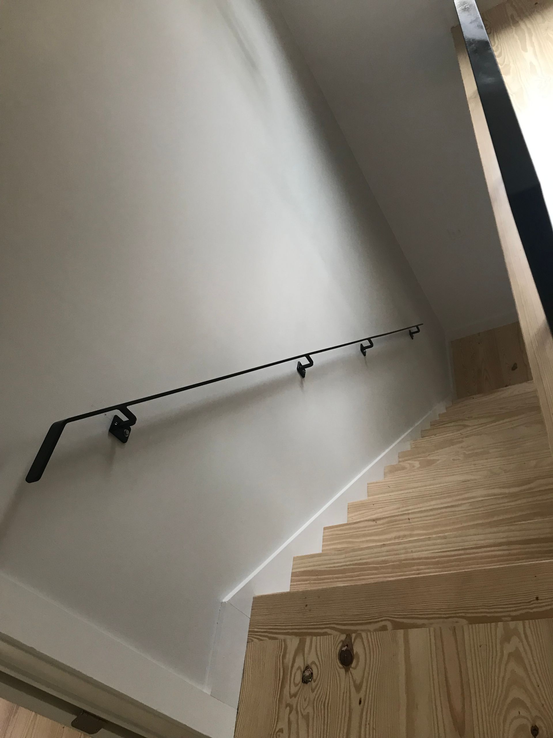 A low-angle view of wooden stairs leading upward, featuring a minimalist black handrail mounted on a plain white wall.