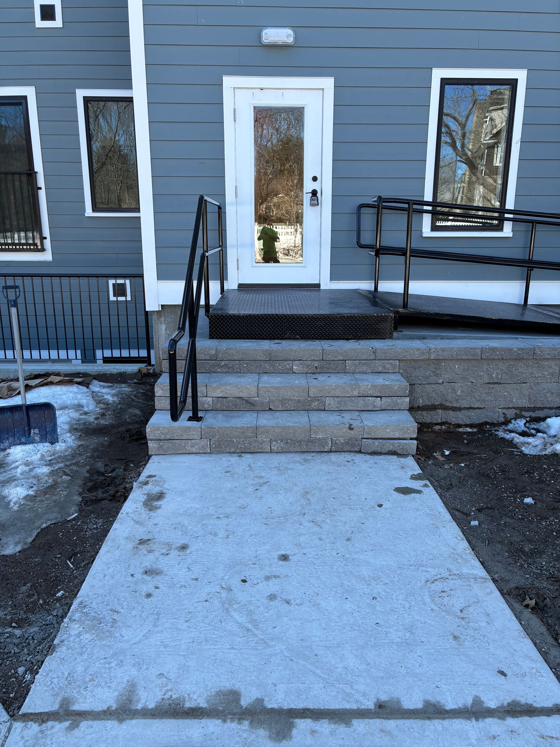 A grey building exterior with a concrete walkway leading to three stone steps, a metal platform, and a white door.