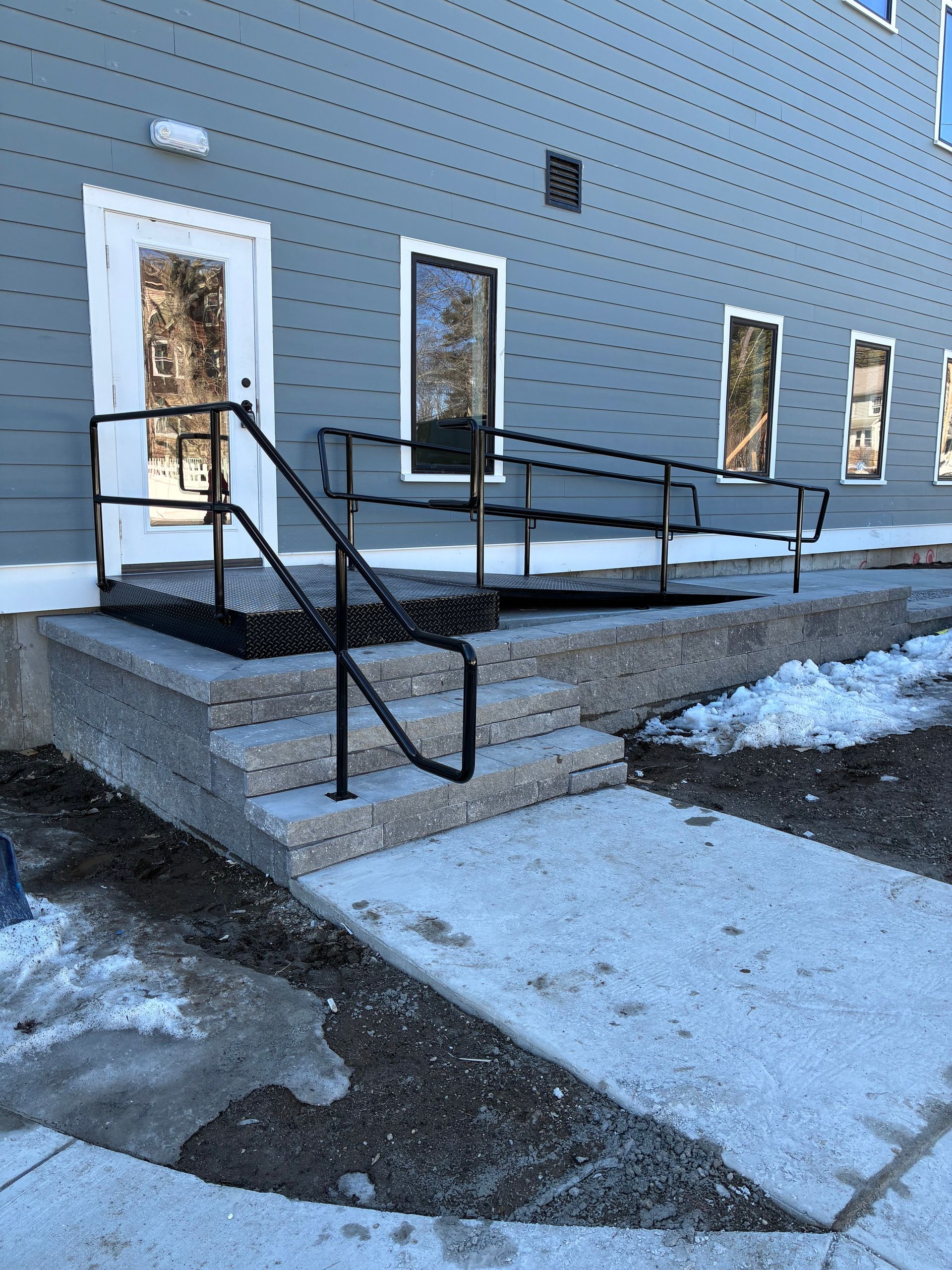 An exterior view of a building with a white door, concrete steps, and a metal wheelchair ramp with black handrails.