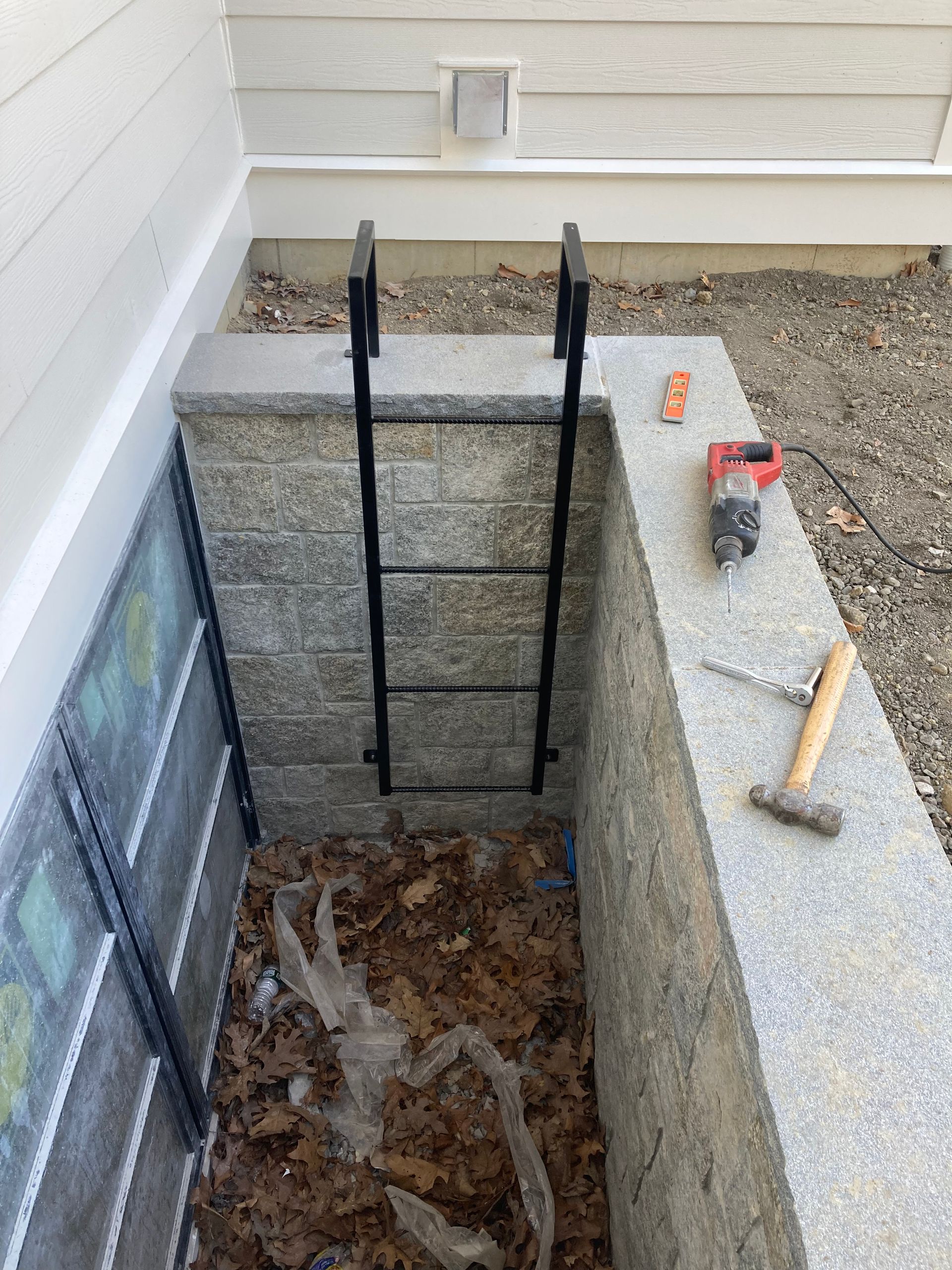 A metal egress window ladder is installed in a stone window well against a house, with construction tools on the ledge.