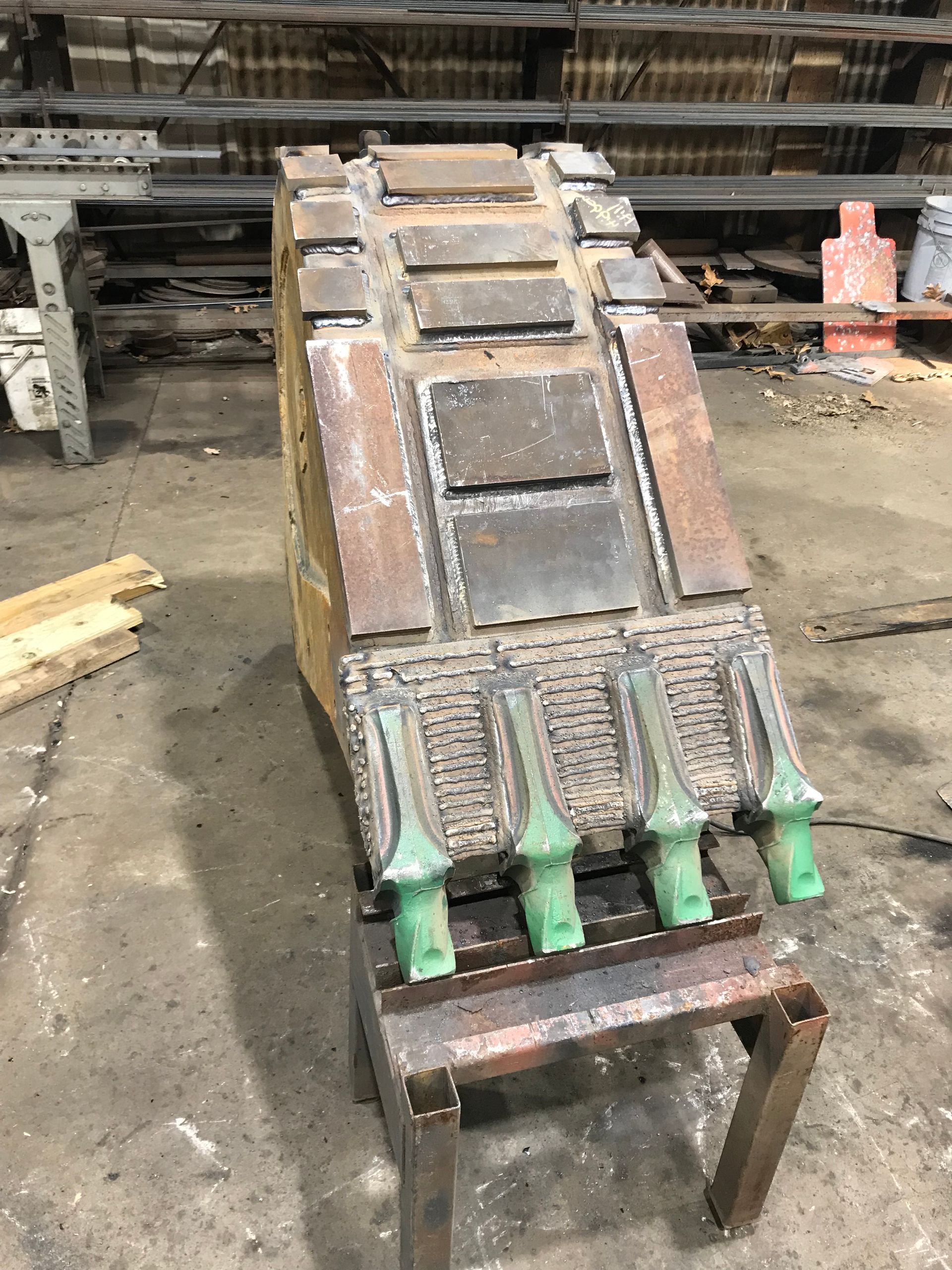 An excavator bucket sits on a metal stand in a workshop, featuring prominent green teeth and reinforced wear plates.