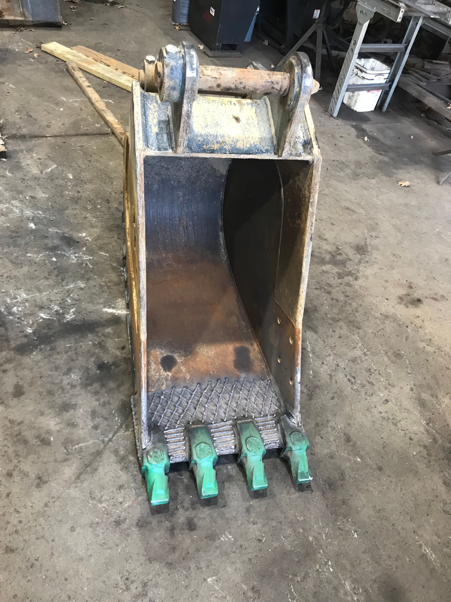 A yellow metal excavator bucket with four green teeth, resting on a concrete shop floor.