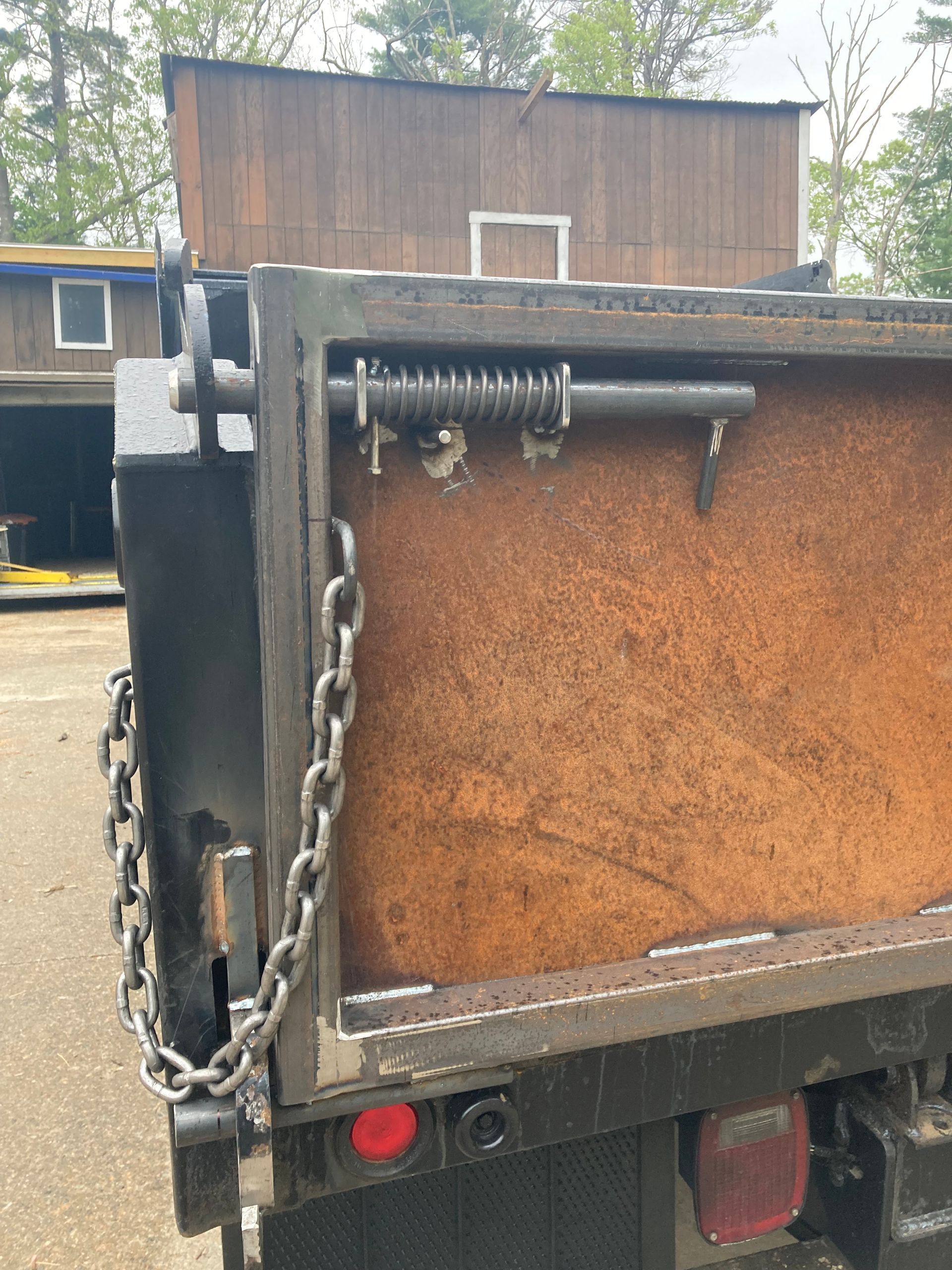 A close-up of a rusted metal dump truck gate featuring a spring-loaded latch mechanism and a heavy-duty steel chain.