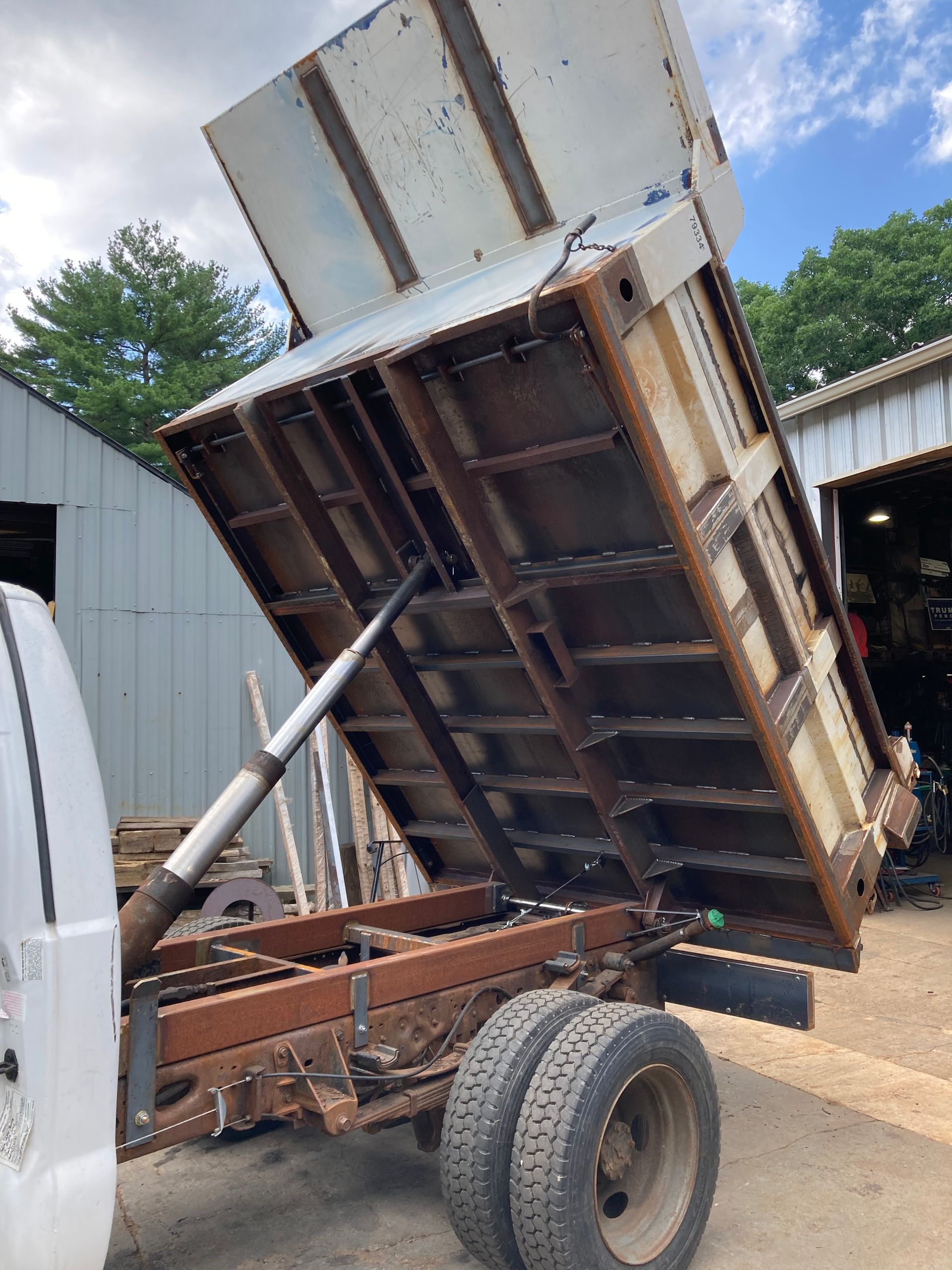 A white truck with its heavy-duty metal dump bed raised, viewed from the side in an outdoor yard.
