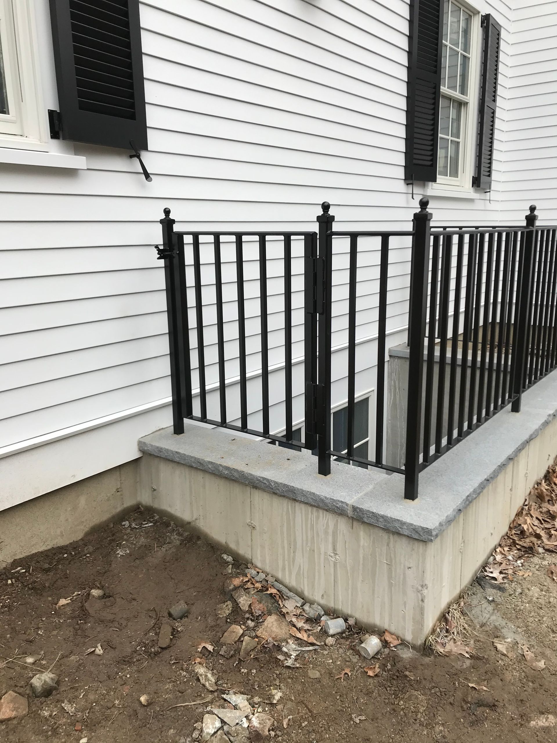 Black metal railing surrounding a concrete window well next to a white house with black shutters and unfinished dirt.