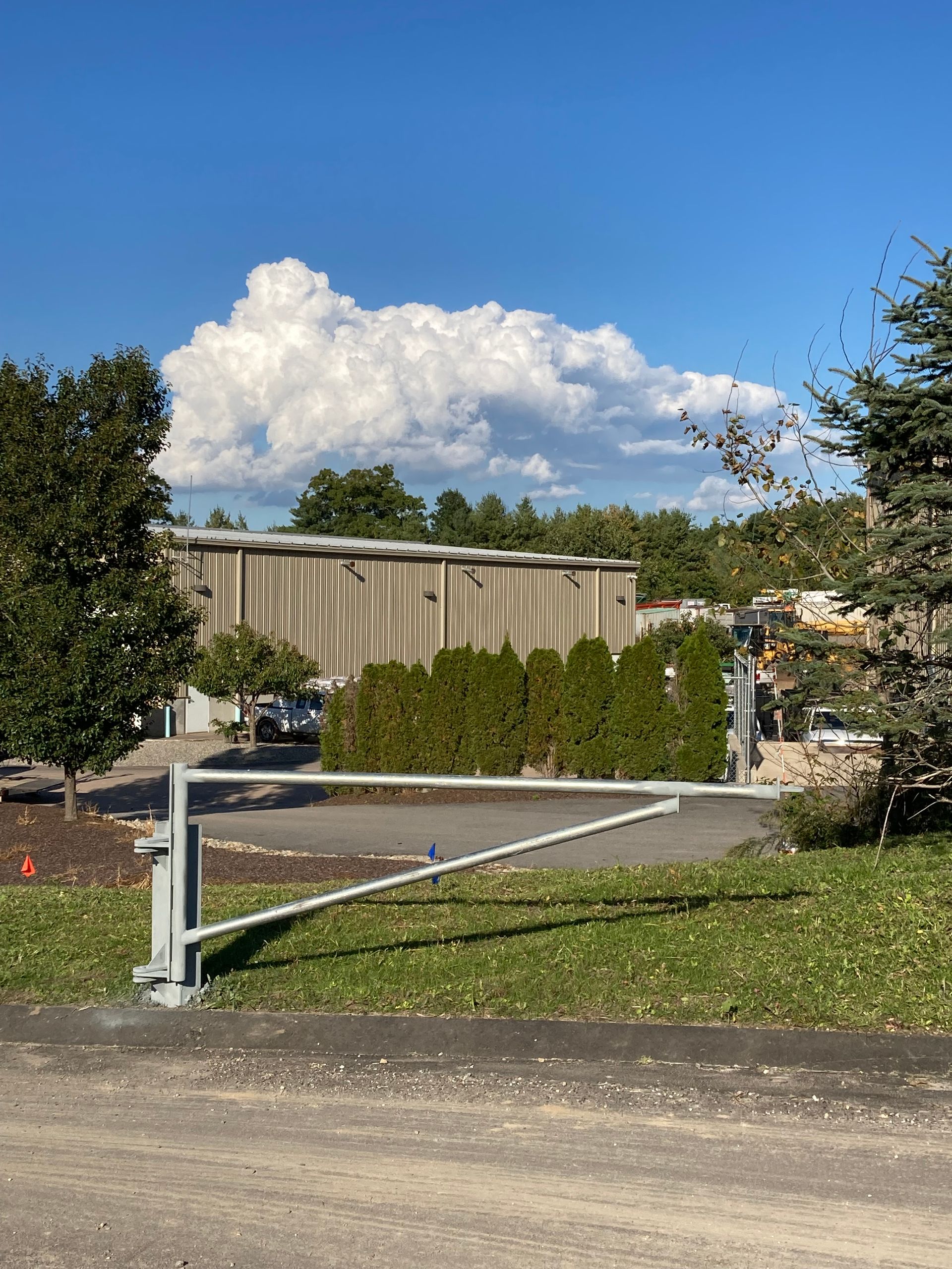 A metal security gate stands on a grassy patch in front of a tan industrial building under a large white cloud.