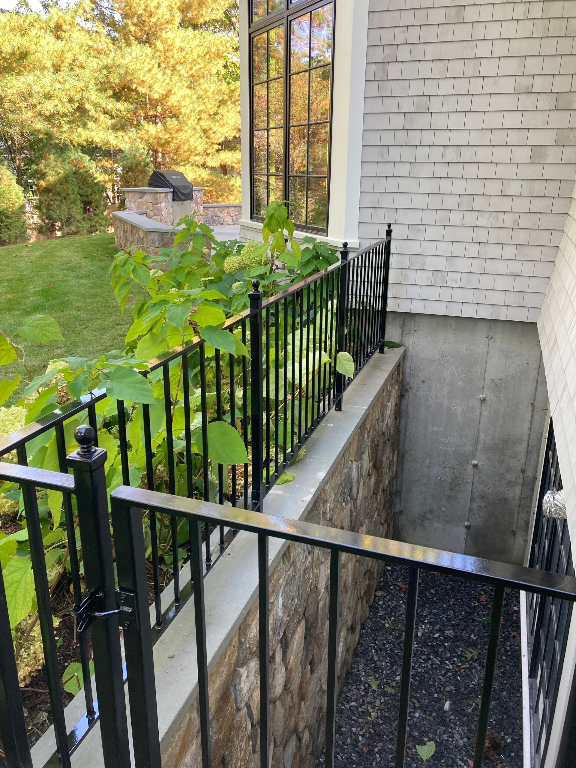 A black metal railing surrounds a stone-walled window well next to a white brick house exterior with green bushes.