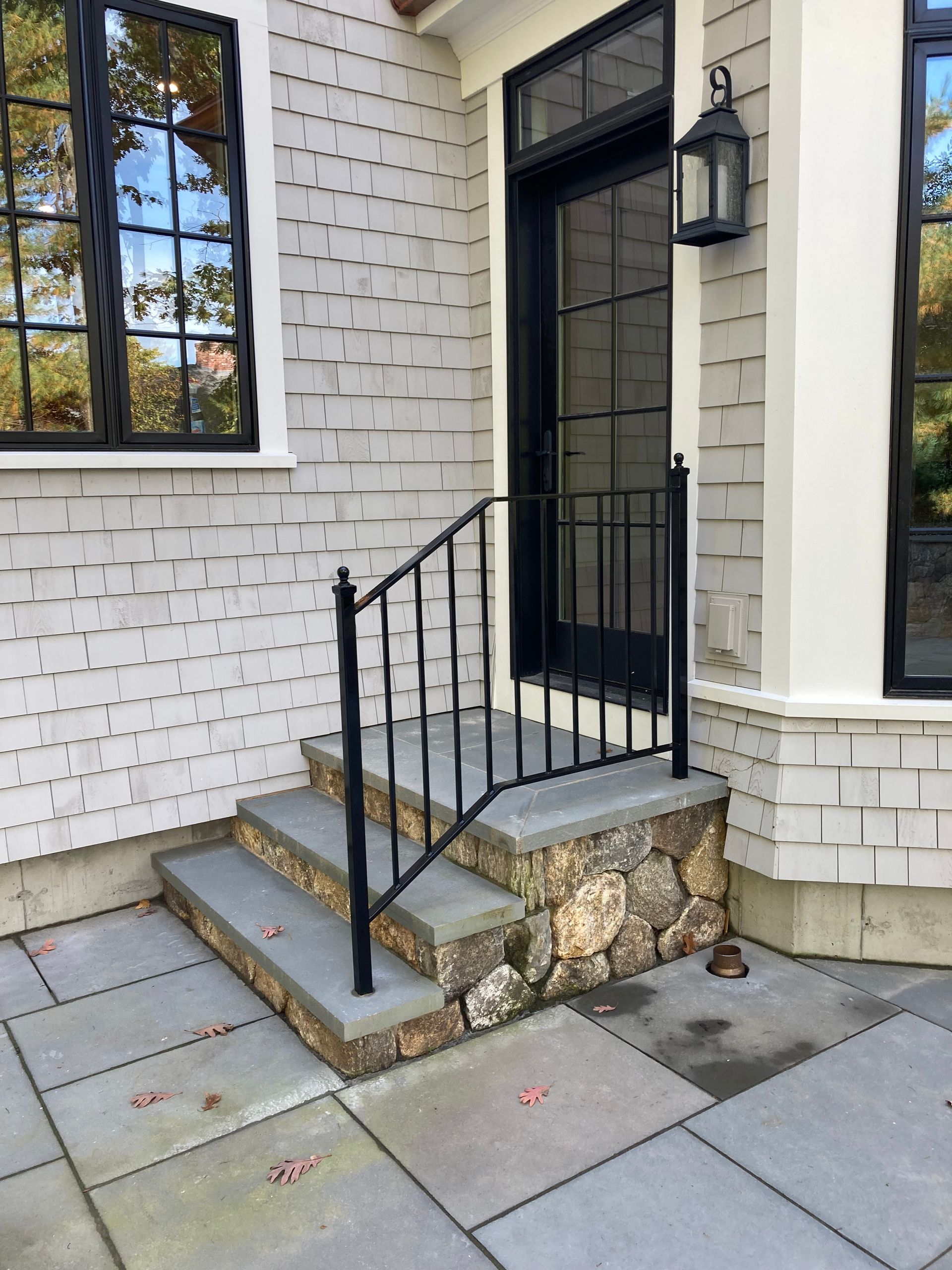 Stone steps with a black metal handrail leading to a dark glass door on a house with light gray wood shingle siding.