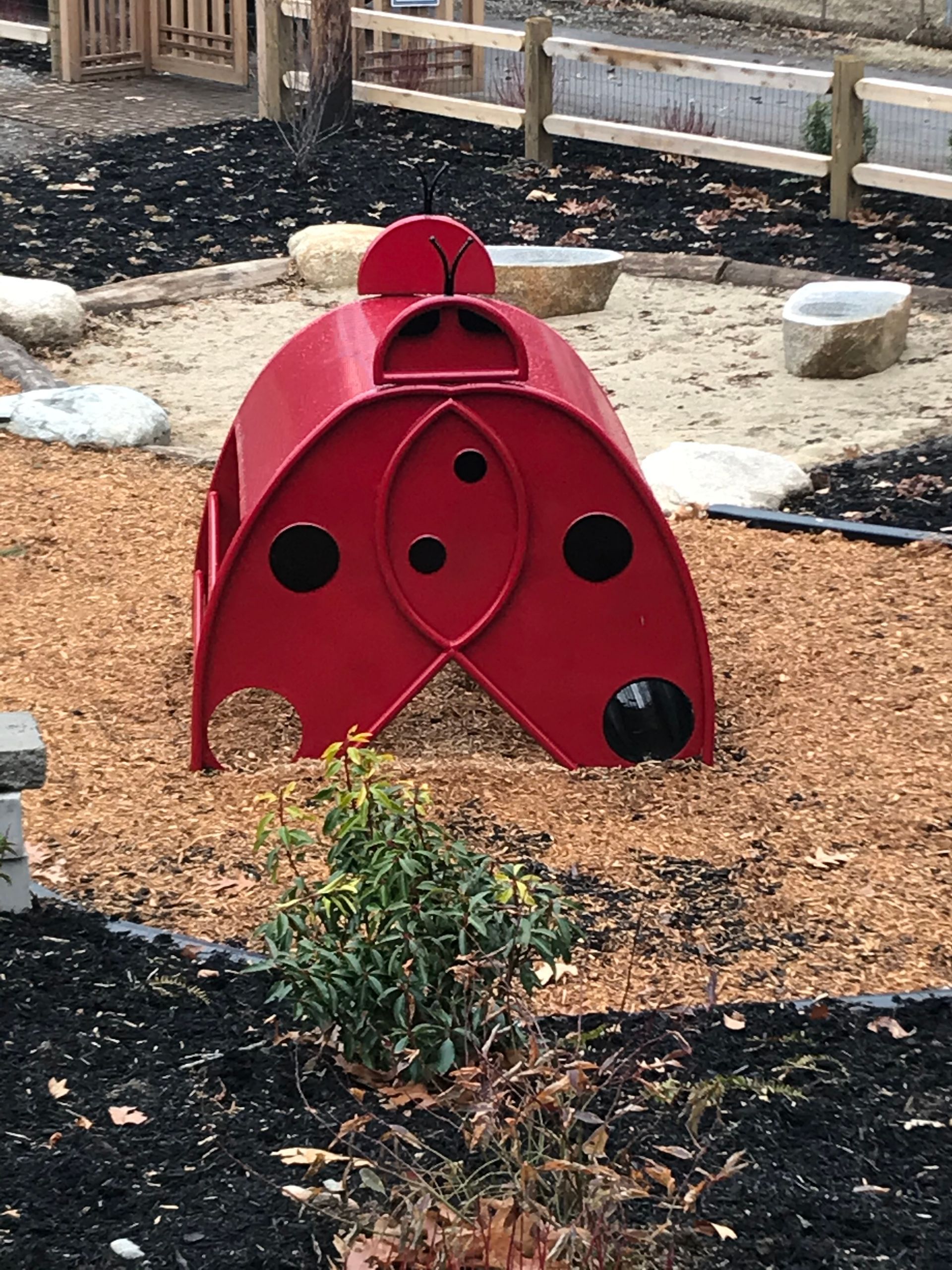 A red, ladybug-shaped play structure with cut-out holes sits on a wood-chipped playground near a sand pit.