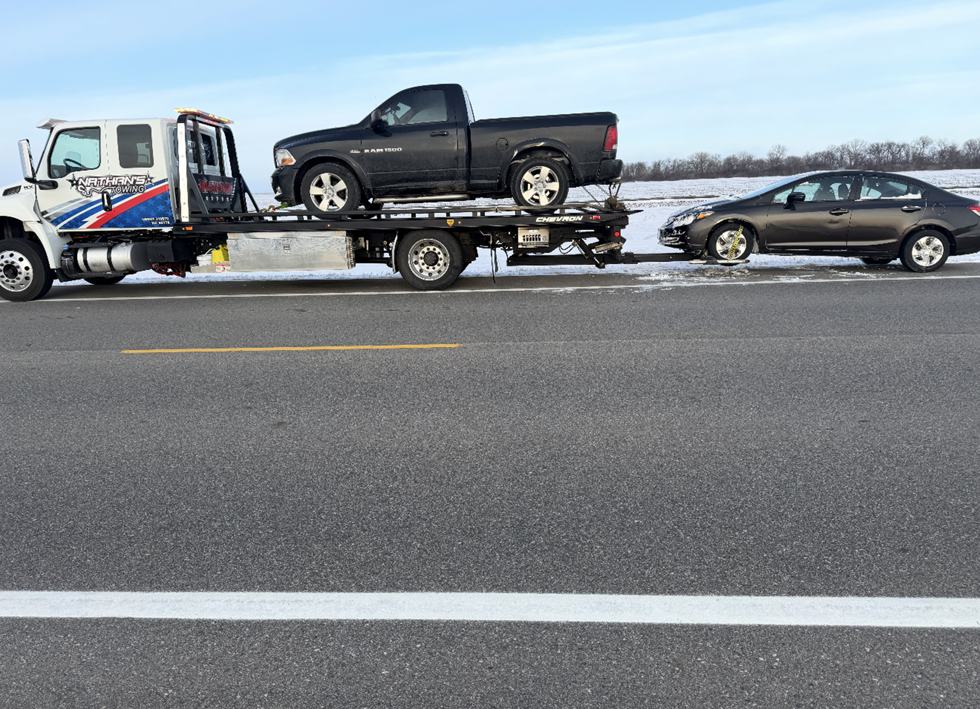 Tow truck hauling a black pickup with a damaged front end, with a black sedan behind on a snowy roadside.