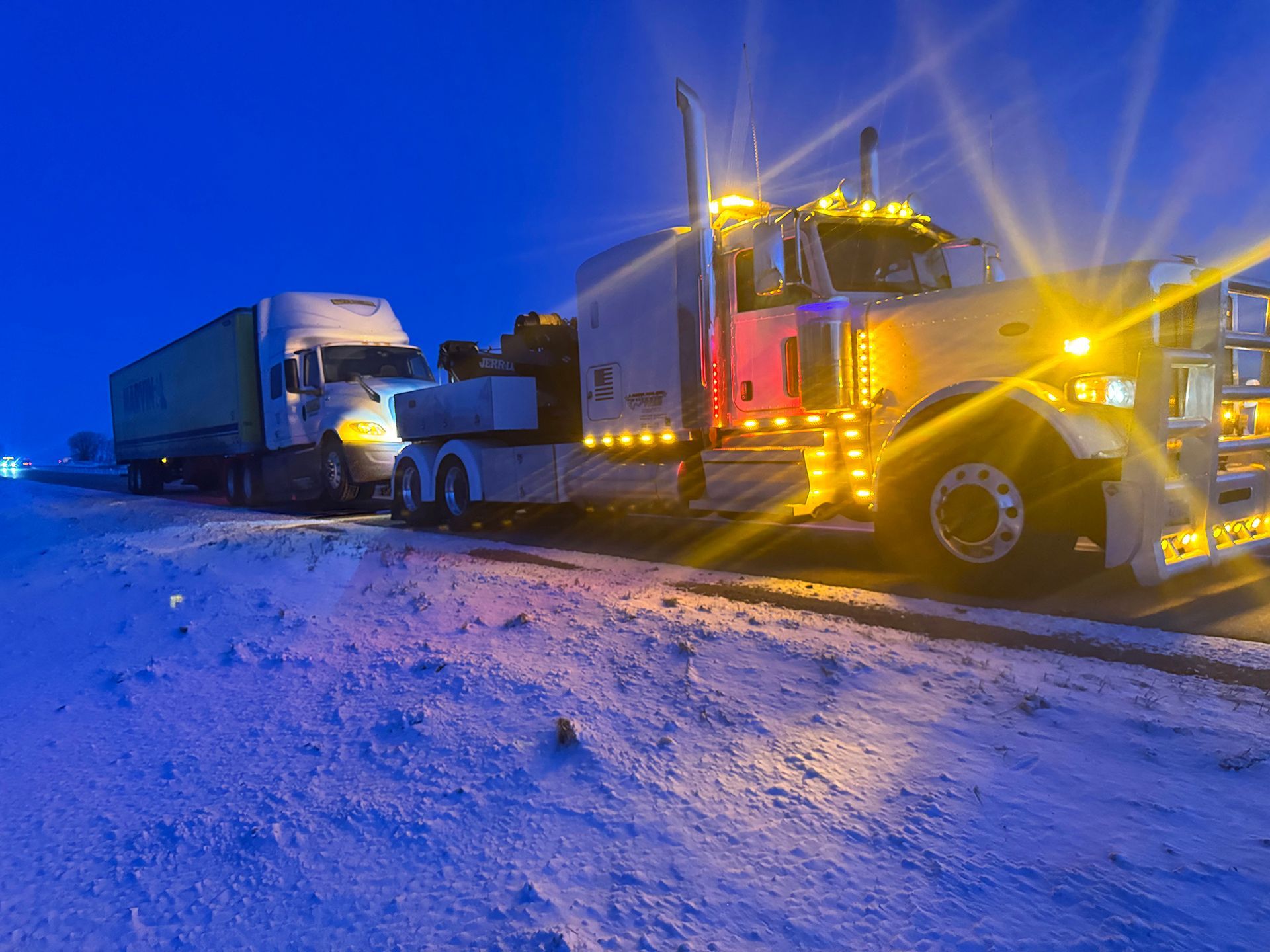 Tow truck towing a semi-truck on a snowy road at dusk, headlights glowing