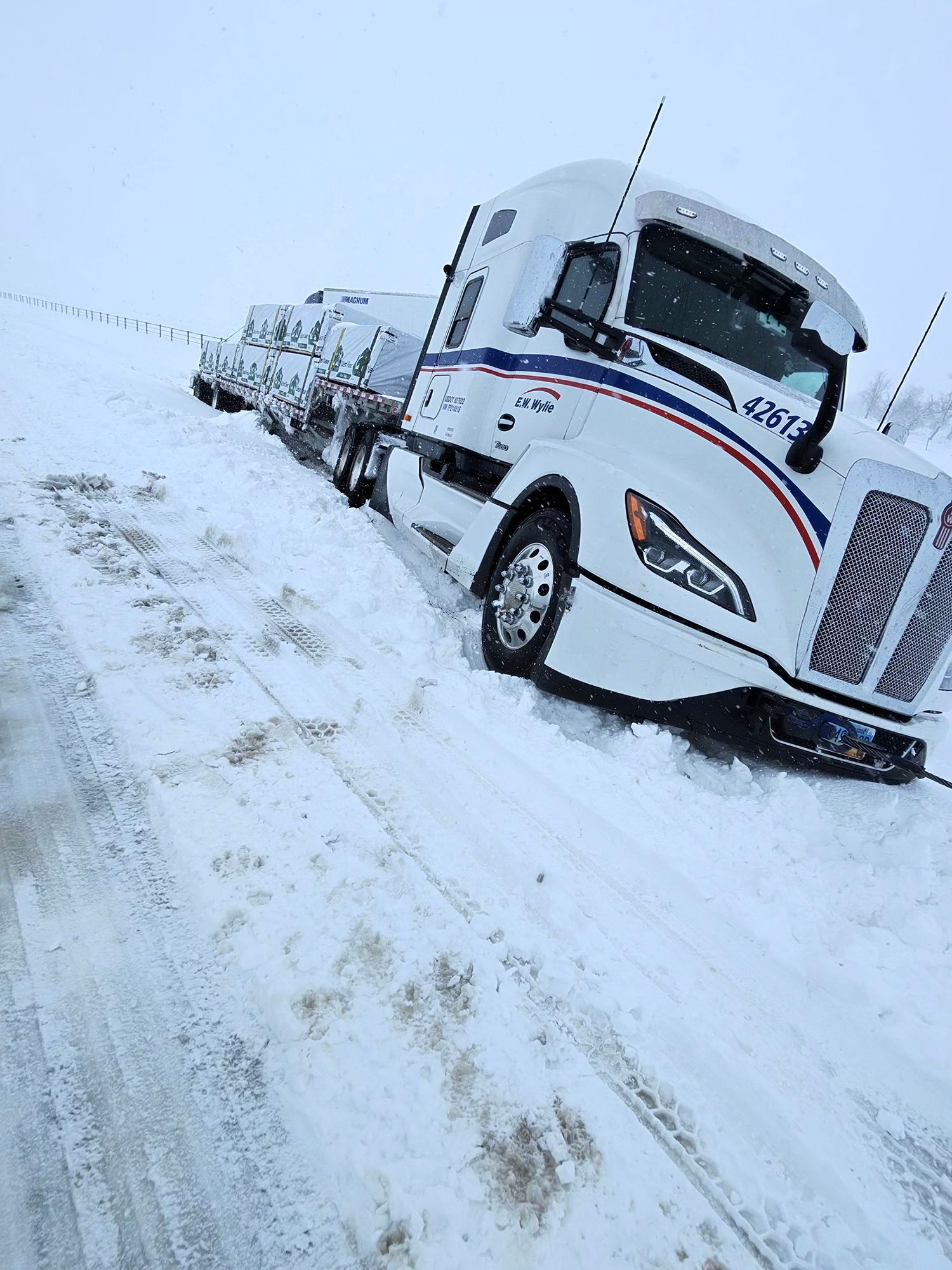 Snow-covered road with a white semi truck and trailer driving in a blizzard