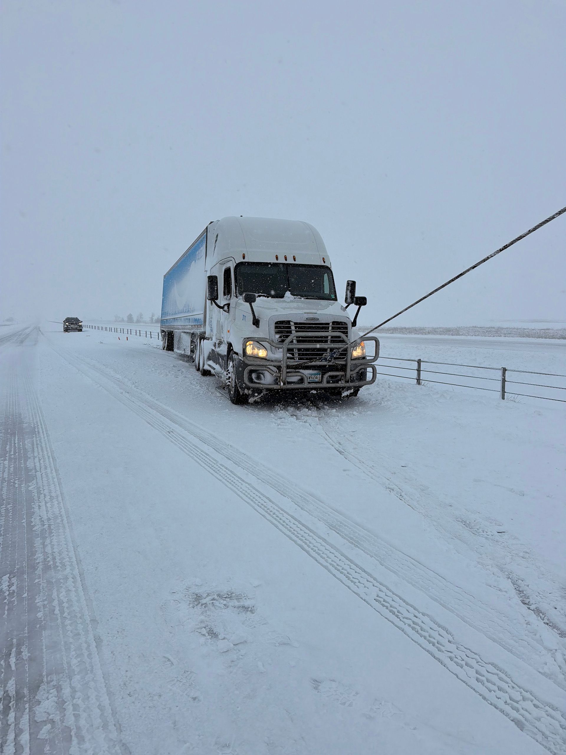 Snow-covered highway with a white semi-truck driving in a blizzard.