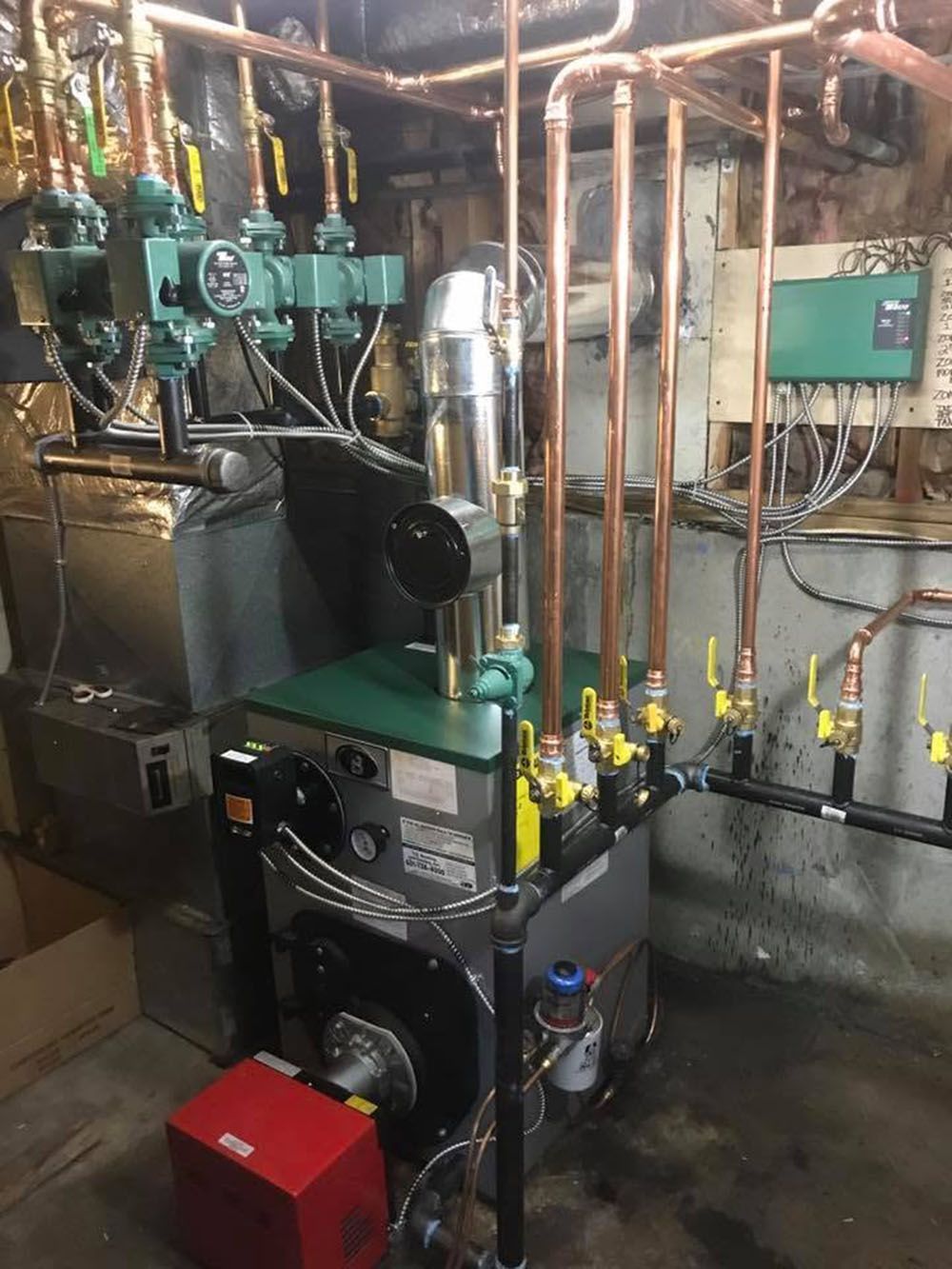 Boiler room with industrial heating equipment, pipes, gauges, and control boxes against a concrete wall.