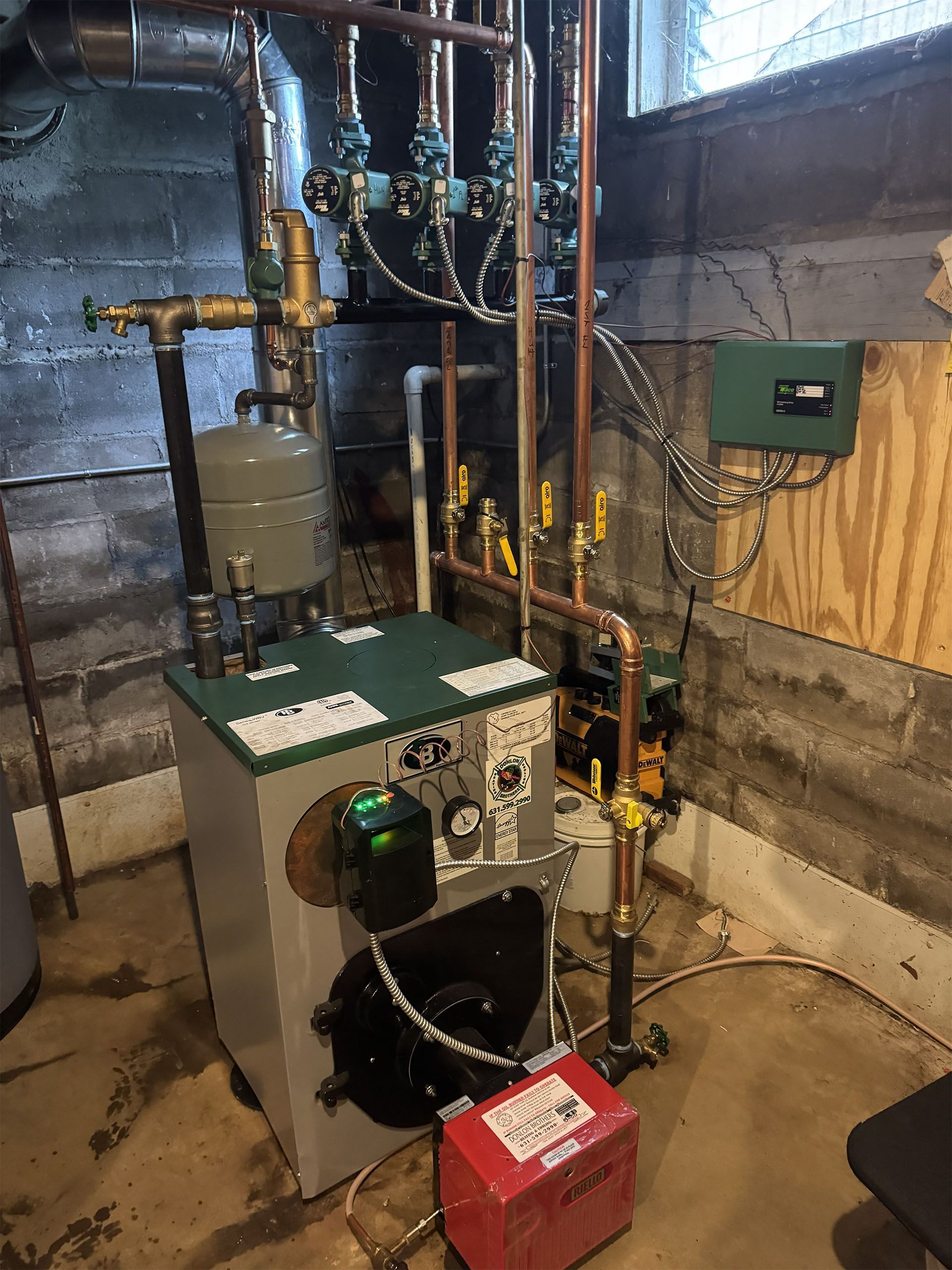 Boiler and piping system in a basement utility room, with a red pump unit in front.