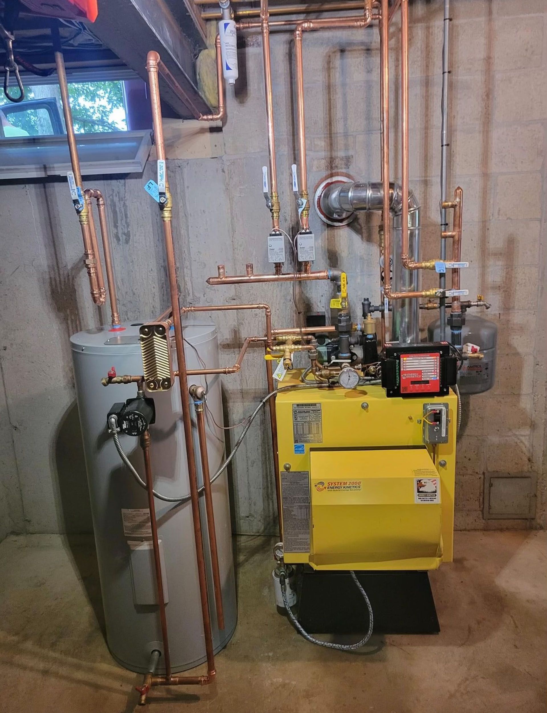 Basement utility room with gray and yellow boilers, copper pipes, and a wall-mounted vent unit.