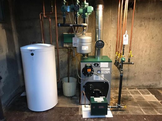 Utility room with a white water heater, boiler equipment, pipes, valves, and a bucket against a concrete wall.