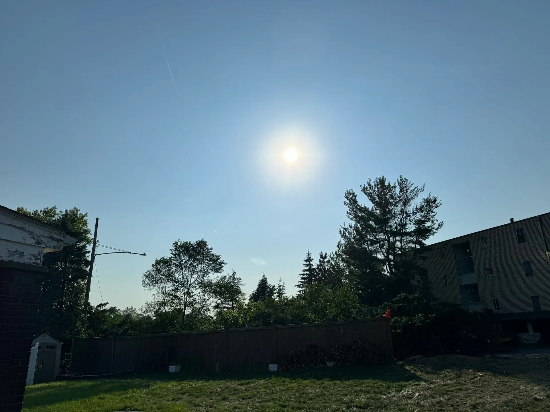 Bright sun in a clear blue sky over trees, a fence, and buildings.