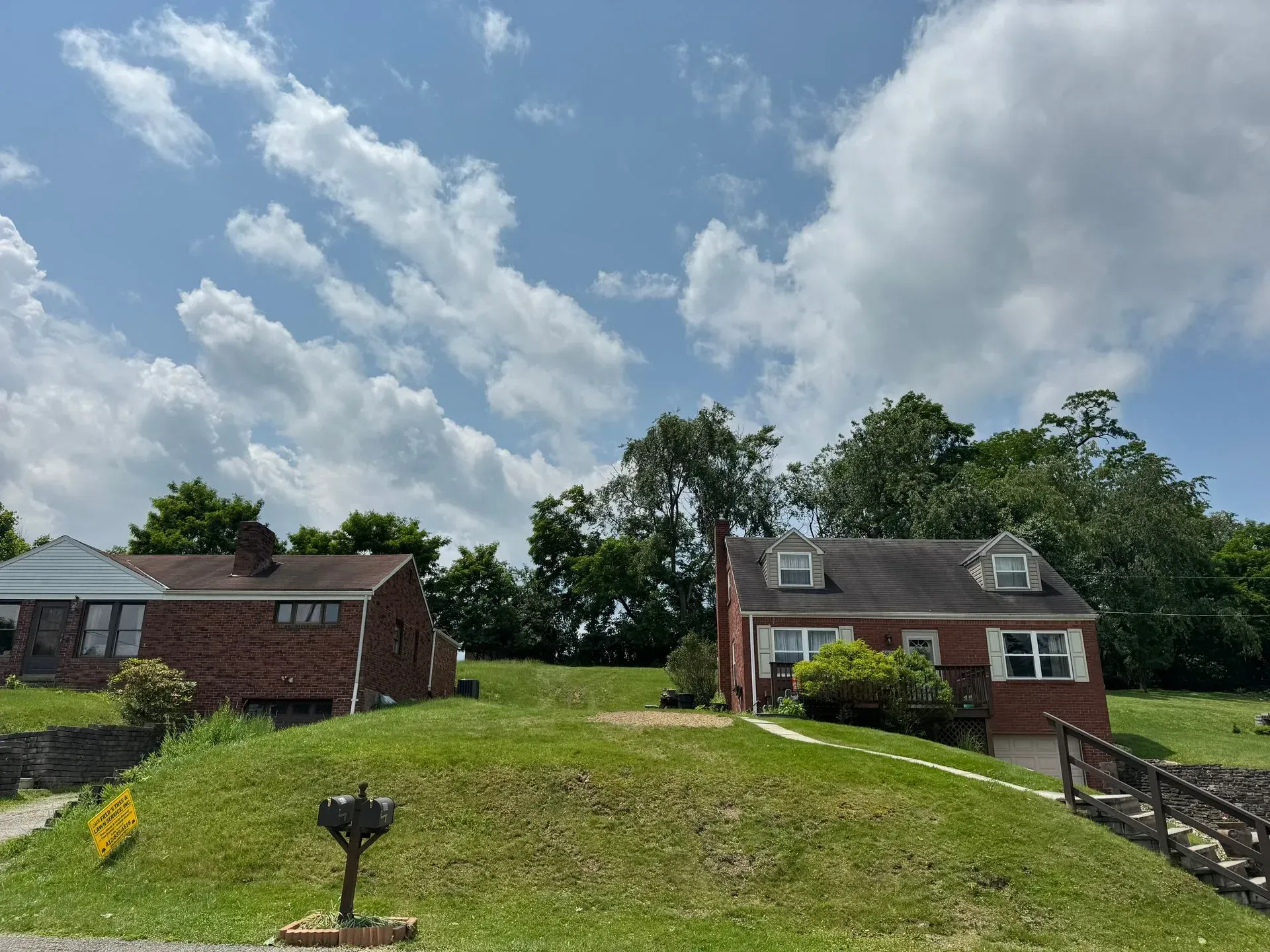 Two houses on a grassy hill with a cloudy blue sky. Trees and a mailbox are also visible.
