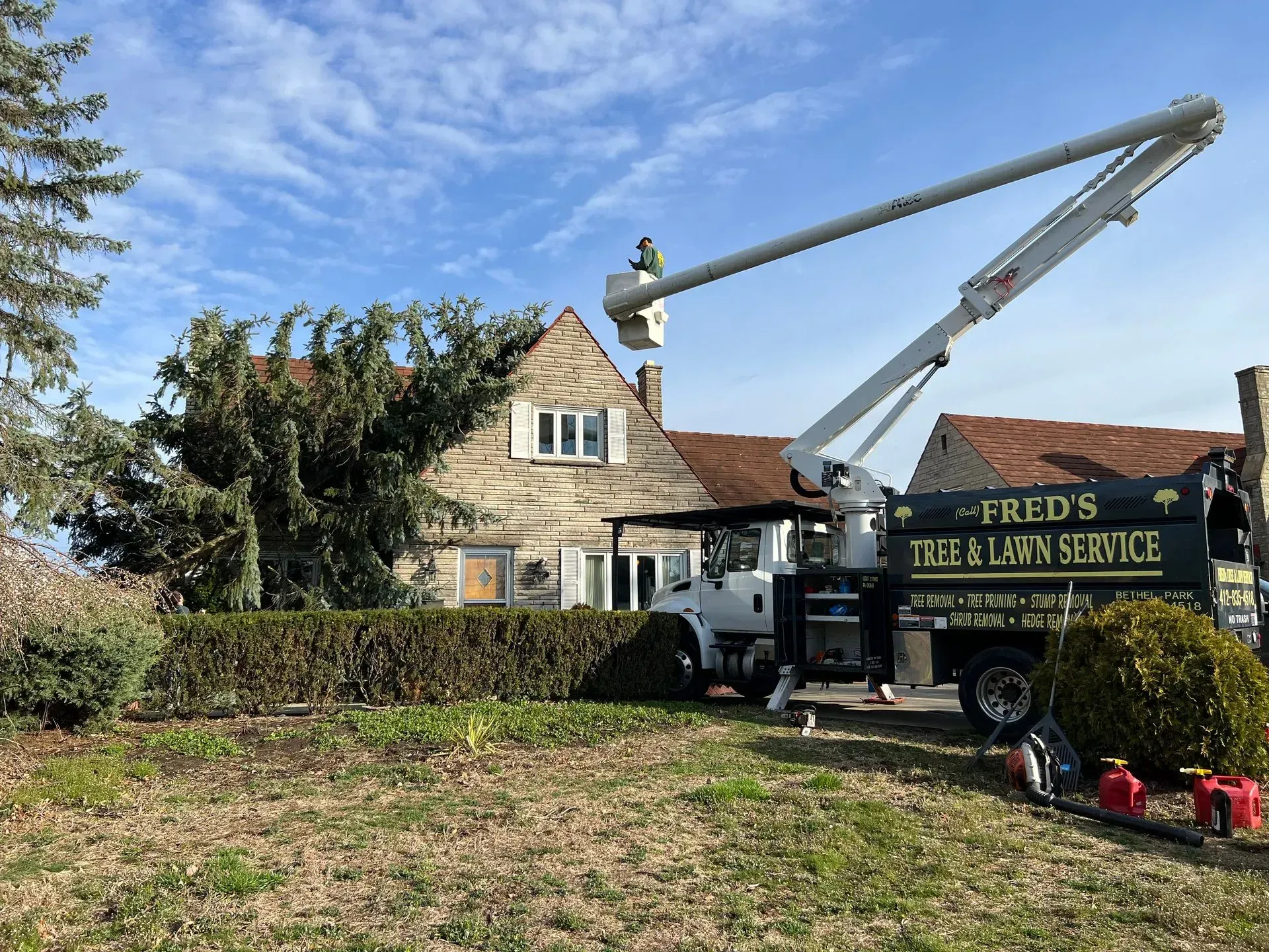 A tree service truck with boom lifts, trimming a tree in front of a house. Sunny day.
